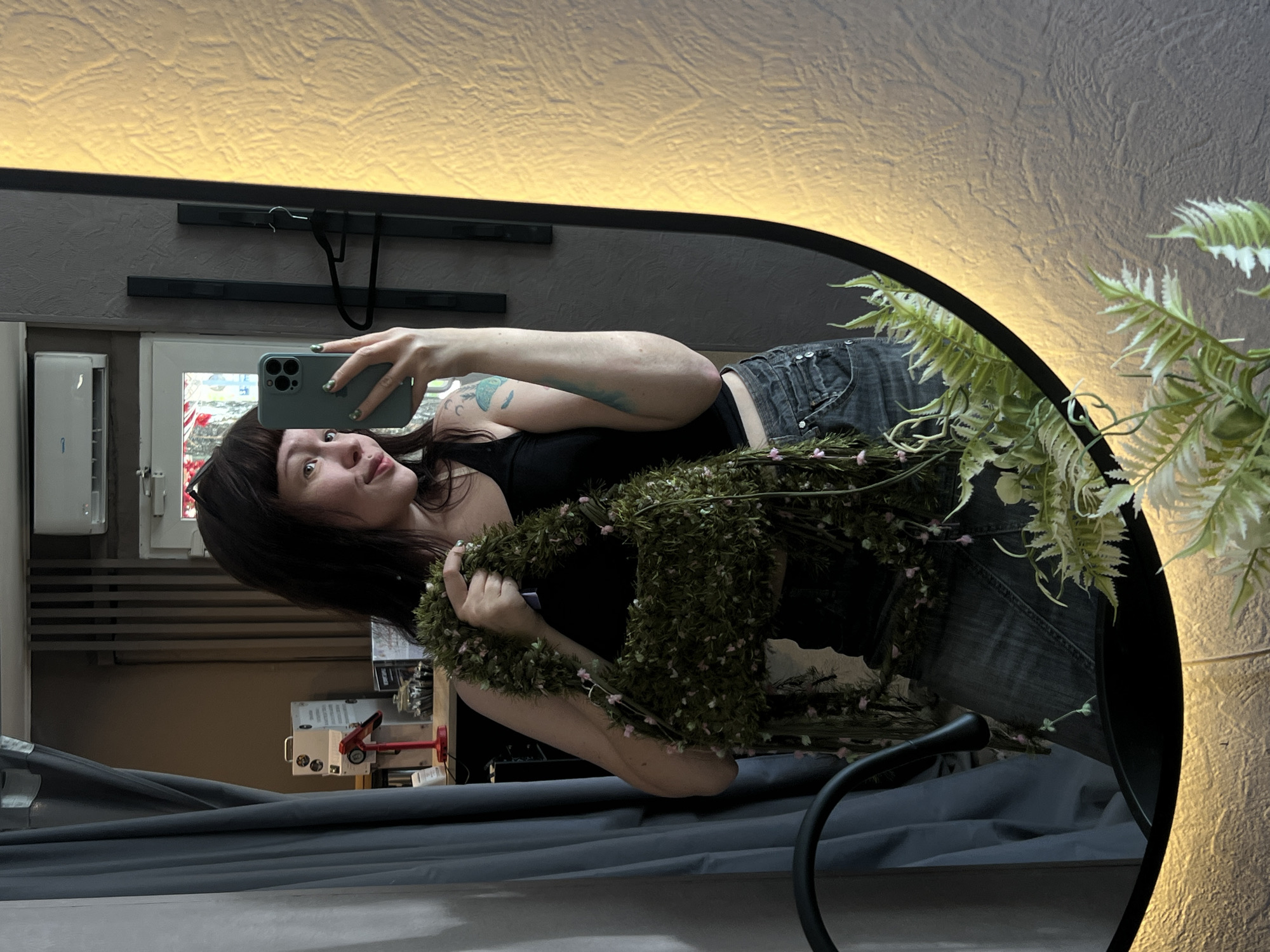 A young woman with dark hair and bangs, wearing a black tank top and denim skirt, is taking a mirror selfie in what appears to be a shop or studio in Chaikovsky, Russia. She is holding a unique, textured green handbag adorned with small pink flowers and dangling greenery. Her right arm, holding a light green smartphone, is visible, as is a tattoo on her left arm. The background shows a dark curtain on the left, a shelf with items including a red object, and a light-colored textured wall on the right. A round mirror with a built-in light frames the scene. A dark chairback is visible in the lower left corner of the frame, and some artificial greenery is in the foreground of the mirror's reflection.