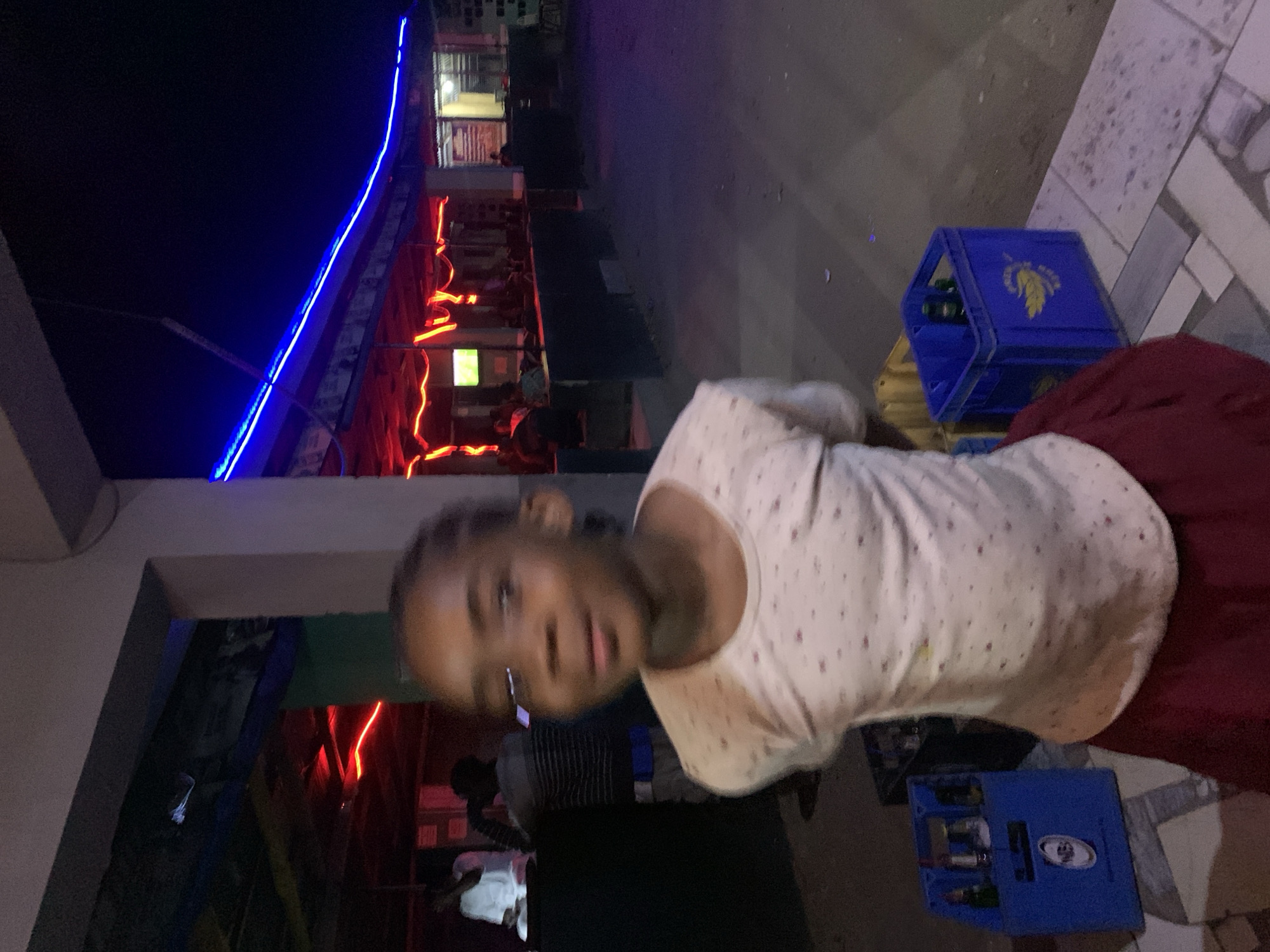 A young girl stands outside a building in the New Stadium area of Nigeria. The building is illuminated with both blue and red neon lights. A blue crate sits on the ground near the girl. People are visible in the background.