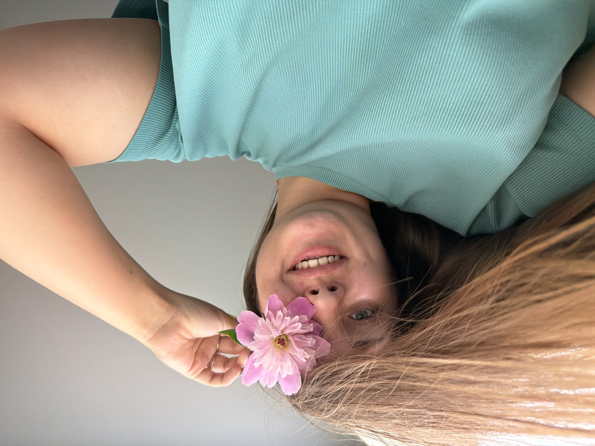 The media file shows a close-up, upside-down, selfie-style shot of a young woman. She is wearing a teal, ribbed top. She is holding a single, light pink peony flower in front of her face, with the petals partially covering her mouth and nose. Her right hand, with a gold ring on her index finger, is visible near the flower. The woman has light skin, light-colored eyes, and her long, light brown hair is spread out around her head. Her expression is a slight smile. The background is a plain, light gray wall. The lighting suggests it is daytime, possibly indoors. There is no visible text. The overall mood is relaxed and playful.