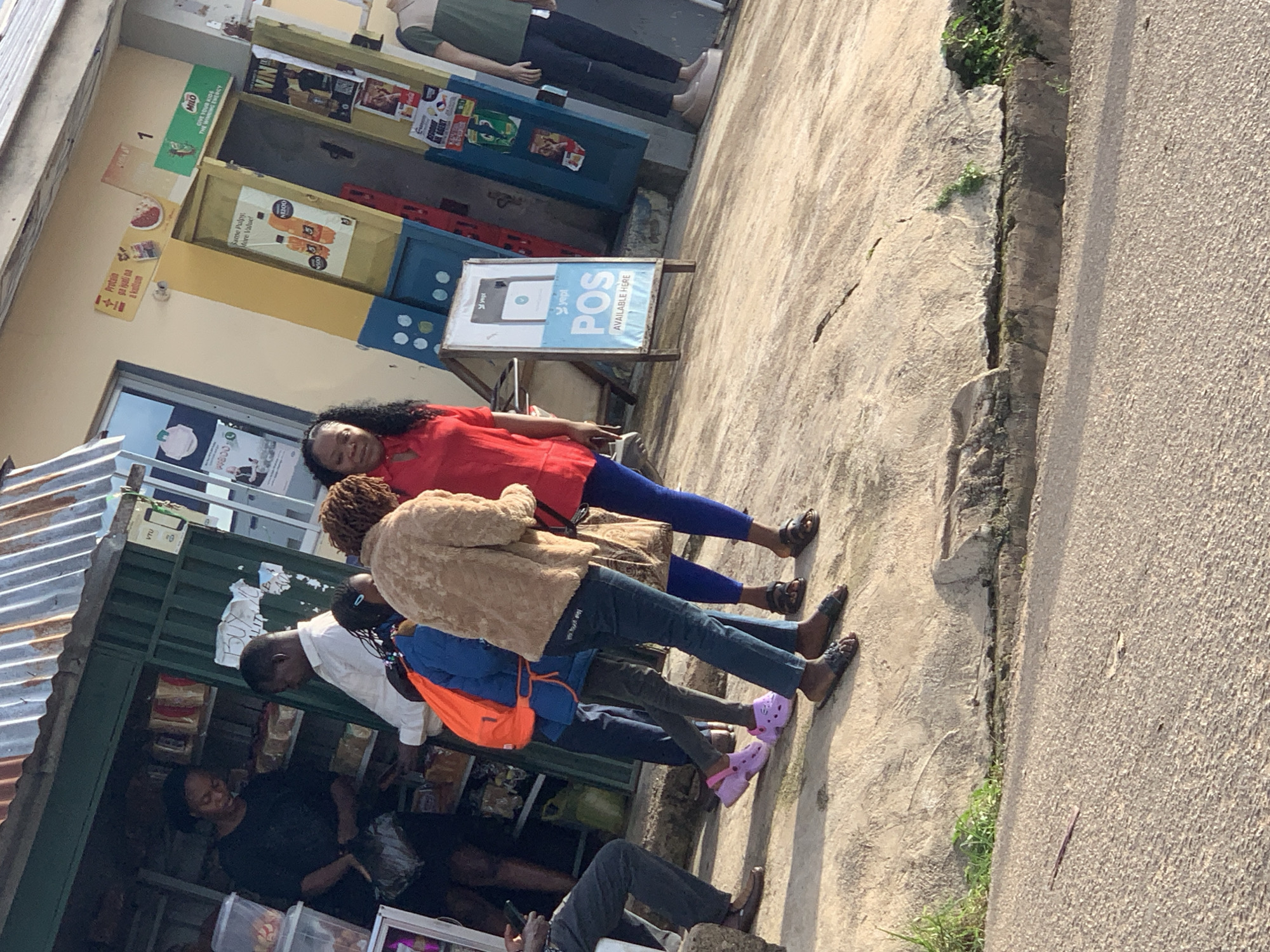 The image shows a street scene in Jos, Nigeria, with several people standing on a sidewalk. The time of day appears to be late afternoon, with the sun casting long shadows, suggesting bright, possibly warm weather.
In the foreground, a woman in a red shirt and blue pants is partially visible, leaning against a structure. Behind her, two more individuals are standing, one wearing a light brown jacket and blue jeans, and the other wearing a white shirt. Further back, other people are seen near a small shop or kiosk, where goods are displayed.
The setting is a street with a paved sidewalk and a road. There are shop fronts with various signs and advertisements visible. One sign advertises "MILO" with the tagline "GIVE YOUR KIDS THE WINNING ENERGY". Another sign displays "POS AVAILABLE HERE" with the logo for "yept". Products like soft drinks and other packaged goods are visible inside the shop. The overall atmosphere seems to be that of a typical street market or commercial area. The people's expressions are not clearly discernible due to the image quality and angle.