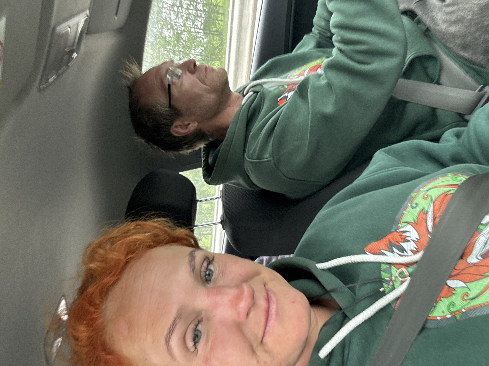 In the interior of a car, a woman with bright orange hair and blue eyes is in the foreground, looking at the camera and smiling. She is wearing a green hoodie with a white drawstring and a design on the front. A seatbelt is visible across her chest. To her right, a man wearing glasses and a similar green hoodie is driving. He is looking forward, out of the car. The car appears to be on a road, with trees visible in the background. The overall scene suggests a couple traveling by car. The location is identified as Tatarstanskie Chelny, Russia.