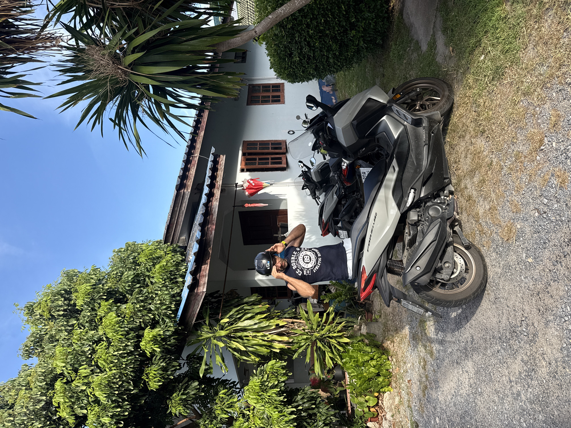 A man stands in front of a house, putting on a black helmet. He is wearing a dark blue sleeveless t-shirt with white text and white shorts. Two motorcycles are parked on a gravel driveway in front of the house. The house is white with a brown roof and two wooden-framed windows. Lush green plants and trees surround the house and driveway. The sky is clear blue. The scene appears to be in a residential area of Ko Samui District, Thailand.