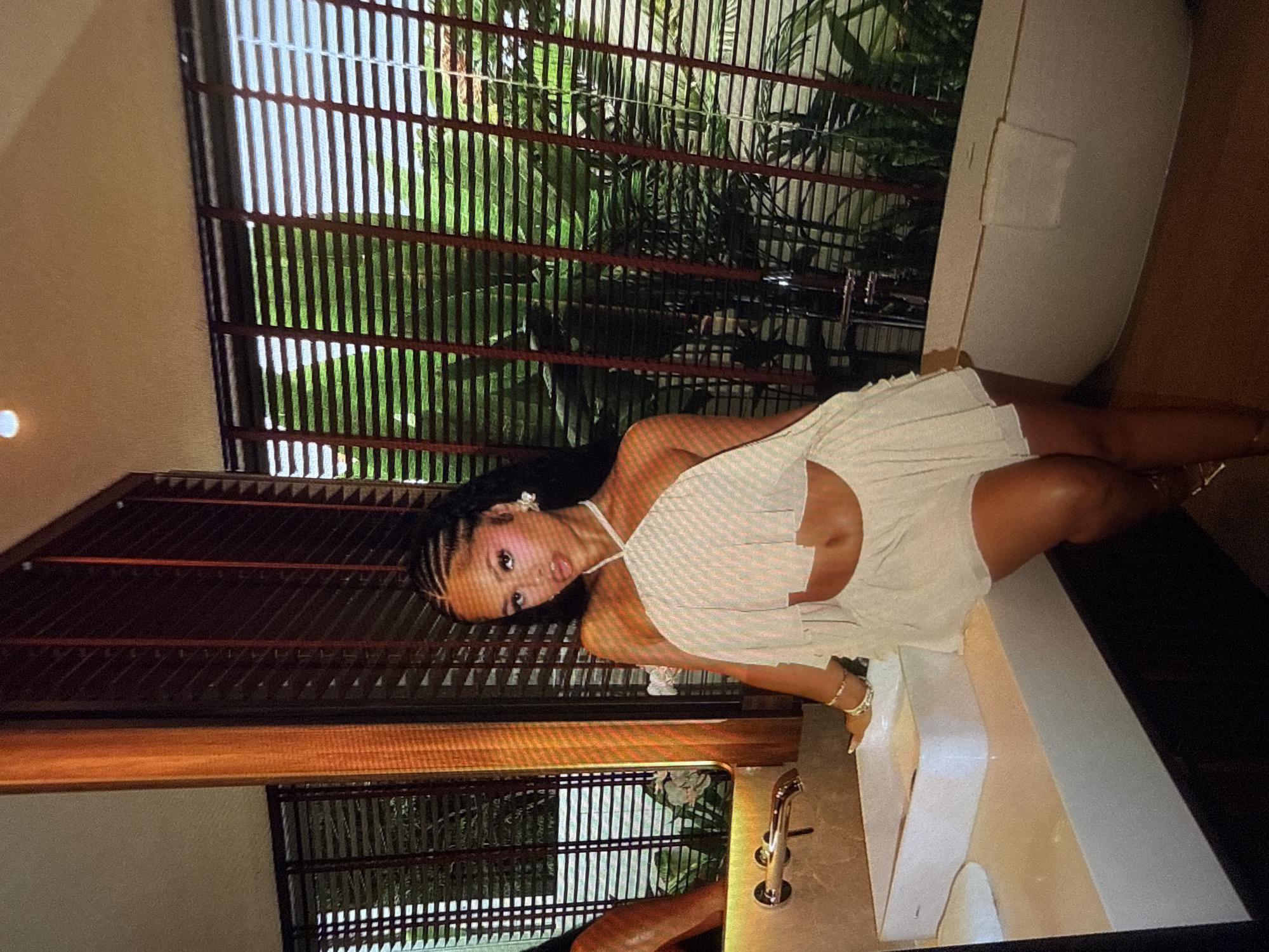A woman is posing in a bathroom. She is wearing a light-colored, two-piece outfit that exposes her midriff and has a halter top with ruffles. Her hair is braided. She is wearing jewelry on her wrists and earrings. She is leaning on a white countertop with a sink. Behind her, there is a bathtub and large windows with blinds, through which lush greenery is visible. The bathroom has a modern aesthetic with a large mirror and a polished finish. The lighting in the room is warm, with a spotlight visible on the ceiling.