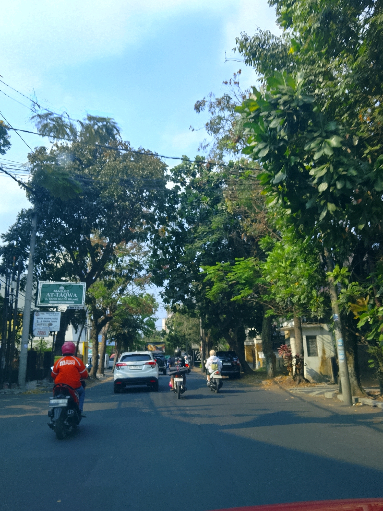The image depicts a street scene in the city of Malang, Indonesia. A person wearing an orange jacket with "m Food" branding and a pink helmet is riding a motorcycle in the foreground. To the right of this motorcycle, a white SUV is driving away from the camera. Further down the road, another person on a motorcycle is visible. The street is lined with trees, and buildings are present on both sides of the road. A green sign with white text, "MASJID AT-TAQWA" and street details, is mounted on a pole on the left side of the image. The sky is blue with some clouds. The lighting suggests it is daytime.