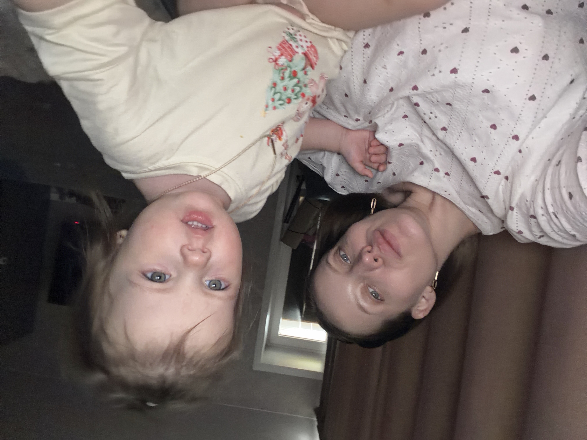 An indoor, medium shot photograph shows two females, an adult and a young child, lying down and looking upwards towards the camera, appearing upside down to the viewer. They are positioned with their heads towards the bottom of the frame.
The child, on the left, has light-colored hair and bright blue eyes. Her mouth is slightly open, revealing some teeth. She is wearing a light-colored (possibly cream or light yellow) short-sleeved top adorned with a festive pattern, which includes what appears to be a decorated Christmas tree with red, green, and white elements, and possibly small animal figures. A thin, light-colored string or cord is visible near her neck.
The adult, on the right, has darker hair pulled back and blue eyes. She has a subtle, pleasant expression, possibly a slight smile. She is wearing a white top with small, scattered red or maroon heart patterns. A gold-colored, rectangular hoop or bar earring is visible in her right ear.
Between the two individuals, a window with a white frame is visible in the background, suggesting some natural light, although the overall lighting in the room appears somewhat dim. To the right of the adult, a brown curtain is visible. The background to the left of the child is dark and indistinct, possibly a floor or furniture.
The image captures a casual, relaxed moment, possibly during playtime or rest, within a home setting. There is no visible text in the image.