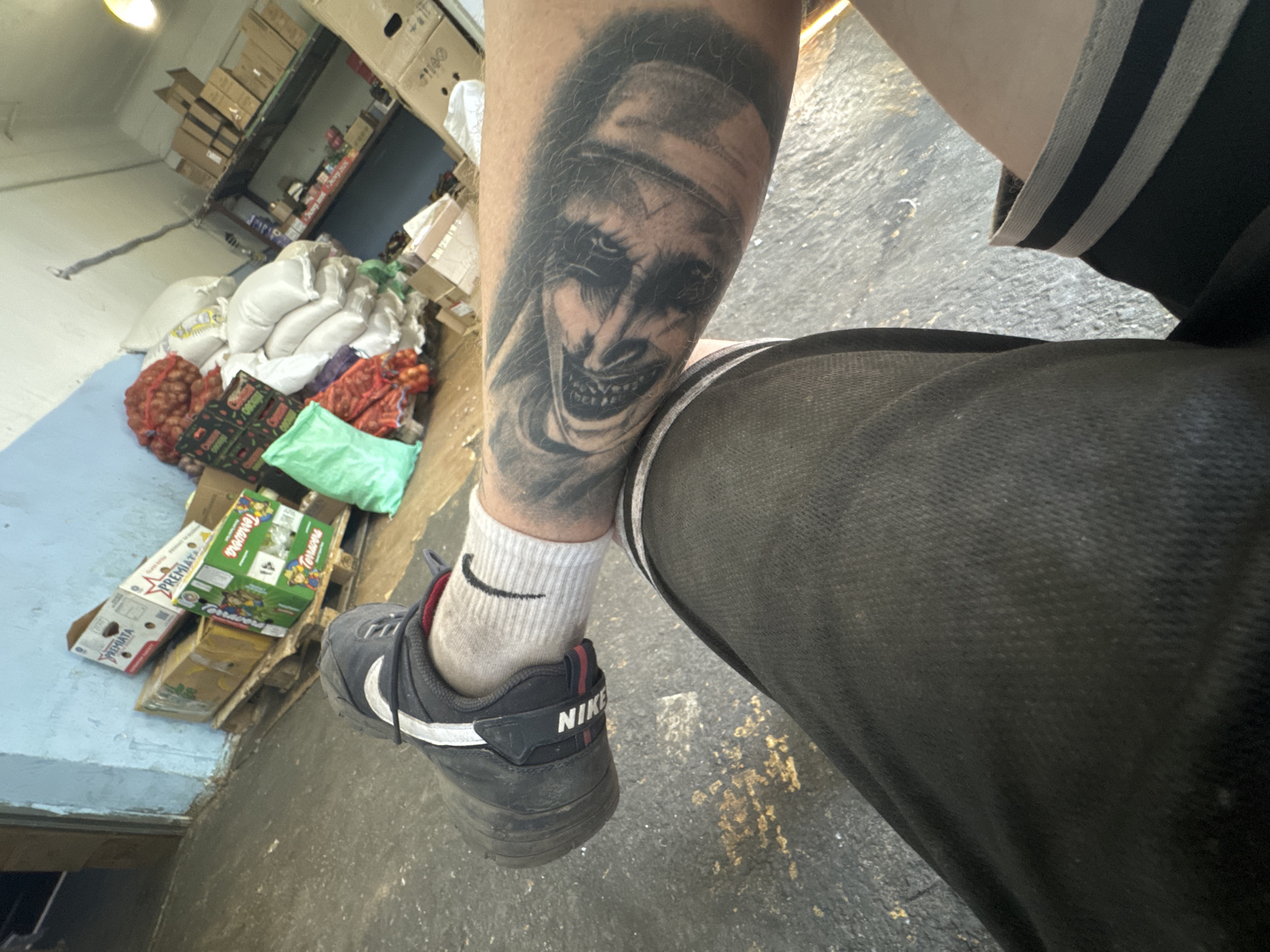The image is a close-up shot of a person's legs and arm, taken from a low angle. The person is wearing black shorts with gray and black stripes at the waistband, white socks, and black Nike sneakers. The person's left arm, visible in the lower right of the image, has a black and gray tattoo of a face. The background of the image appears to be an indoor storage area or warehouse, with concrete flooring and a light blue painted wall. Various boxes and bags of produce, including what appear to be onions and carrots, are stacked on pallets and shelves. The lighting is dim, suggesting an interior setting with artificial light sources.