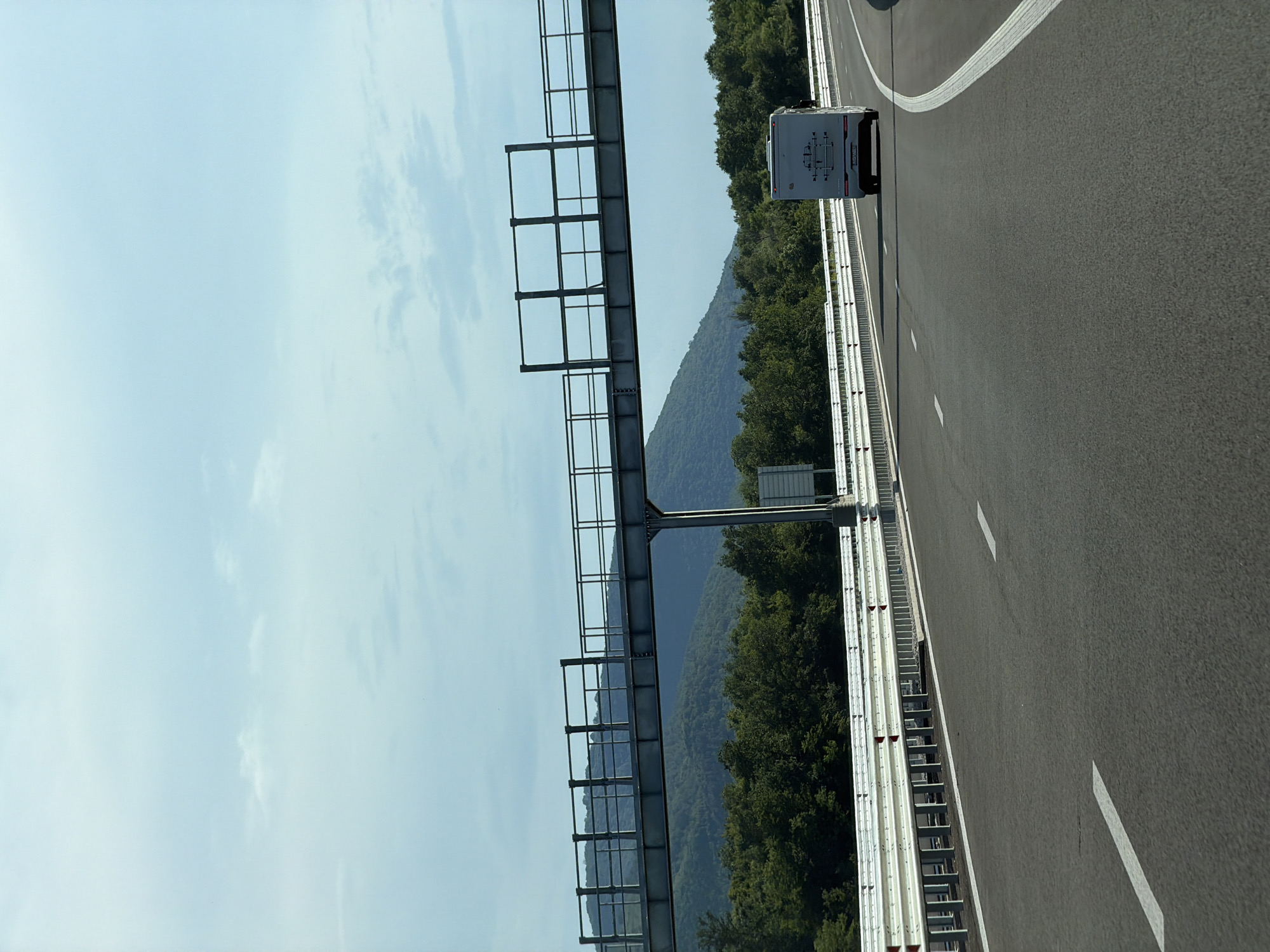 The image shows a highway with a white motorhome driving on it. The highway has a metal guardrail on the side and a large metal structure overhead, possibly for road signs or lighting. In the background, there are green hills covered in trees and a blue sky with some clouds. The scene appears to be in a rural area, and while the image is presented as being in the city of Peshterna, Bulgaria, there are no specific landmarks or details within the image to confirm this location.