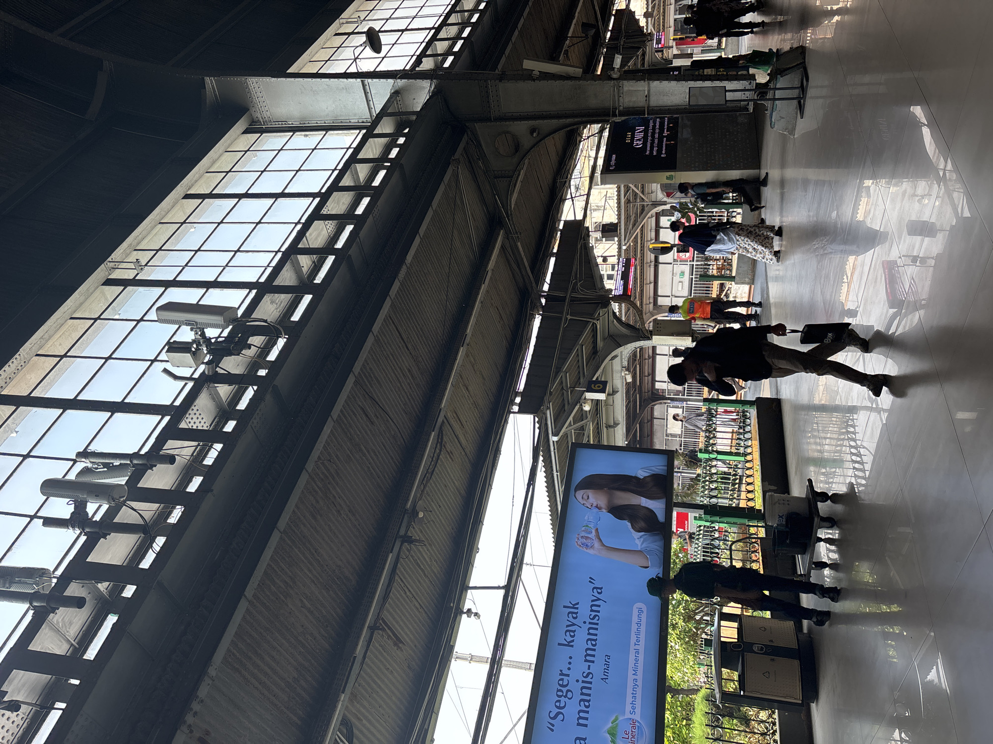 The image shows the interior of a train station, likelyGambir Station in West Jakarta, Indonesia, during the day. Sunlight streams in through large, gridded windows in the ceiling and upper walls, illuminating a polished, reflective floor. Overhead, a complex network of metal beams, corrugated metal roofing, and suspended lighting fixtures creates a sense of industrial architecture.
In the foreground, several individuals are present. A man in a dark uniform with a hat walks away from the camera, pulling a cleaning cart with buckets. Another man in casual clothing walks towards the camera, carrying a black shopping bag. Other figures, some in uniforms and some in civilian attire, are scattered throughout the station, some walking, others standing.
A prominent digital billboard displays an advertisement for "Le Minerale" bottled water, featuring a woman drinking from a bottle. Another smaller digital display is visible further down the platform. The environment suggests a busy public transportation hub, with elements of infrastructure and commercial advertising.