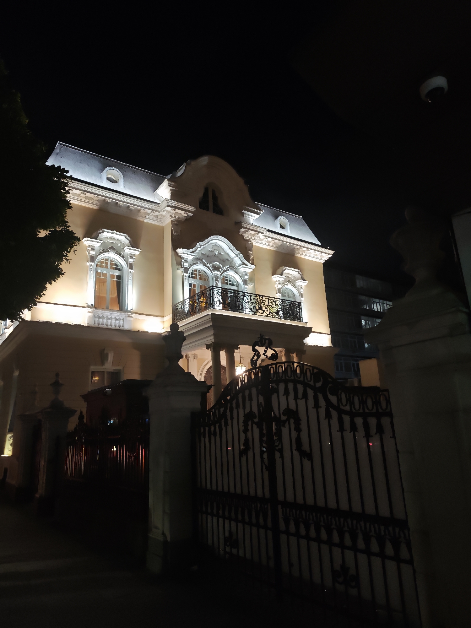 The image shows a three-story building illuminated at night. The building is light yellow with white trim around the windows and roof. It has a balcony on the second floor with black wrought iron railings. A wrought iron gate stands in front of the building, flanked by two white pillars. The sky is dark, and a security camera is visible in the upper right corner. A dark building can be seen behind the yellow one.
The location is likely in the city of Sofia, Bulgaria.