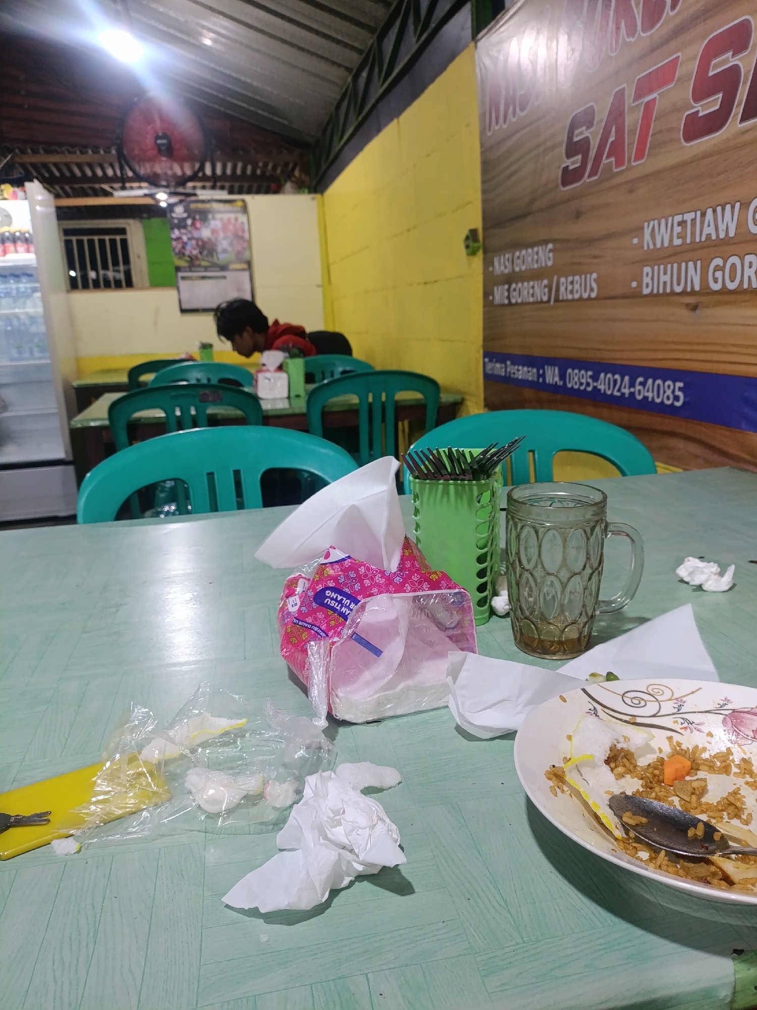 The image depicts the interior of a humble Indonesian eatery, possibly located in Jonggol, Indonesia. The scene captures a table setting that appears to have been recently used, suggesting a meal has just concluded or is in progress.
In the foreground, a green plastic table is laden with remnants of a meal. A white plate contains leftover fried rice with pieces of carrot and what appears to be some form of cracker or fried dough. A silver spoon rests in the plate. To the left, crumpled white napkins and plastic wrappers are scattered, alongside a yellow object that might be a placemat or a tray. A packet of pink and white napkins sits prominently on the table. Beside the napkins, a green plastic container holds several metal forks. A clear glass mug, partially filled with a brown liquid, is positioned next to the forks.
In the background, several teal plastic chairs are visible, some occupied by what appear to be patrons. One individual, possibly a young male wearing a red shirt, is seated and looking downwards, engaged in their meal or activity. The walls of the establishment are painted yellow, and a large banner with text in Indonesian, likely advertising menu items such as "NASI GORENG," "KWETIAW," and "BIHUN GORENG," is affixed to the wall. A ceiling fan is mounted overhead. A refrigerator unit is partially visible on the far left. The lighting is a mix of overhead artificial lights, creating a somewhat dim and casual atmosphere. The overall impression is that of a local, no-frills dining establishment.