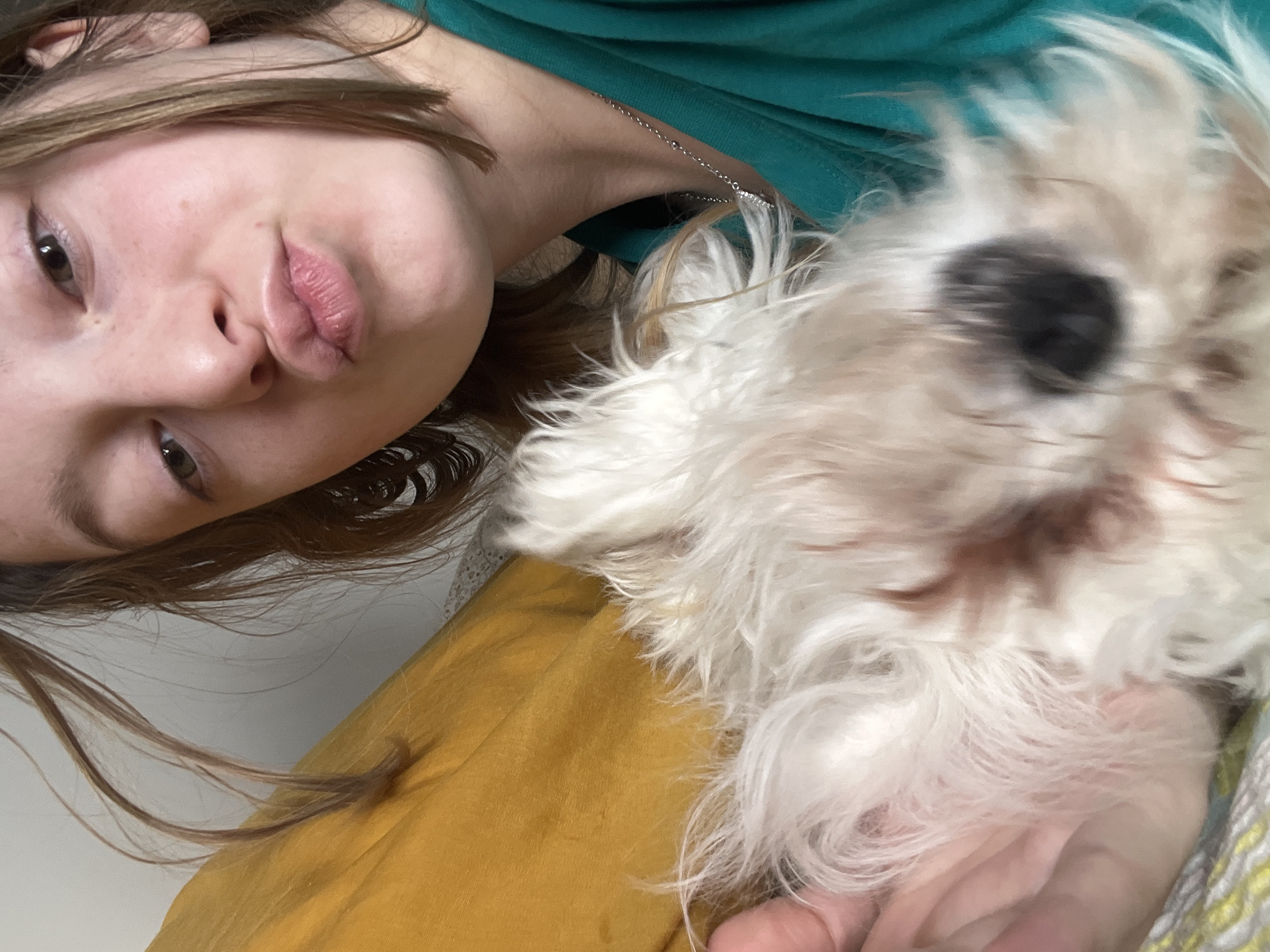 A selfie-style photograph shows a young woman with light brown hair and fair skin. Her face is tilted towards the camera, and she is making a slight kissing expression. She is wearing a green top and a silver necklace. A small white dog with a dark nose and fluffy fur is positioned to the right of her face. The dog appears to be slightly out of focus. Below them is a yellow surface. The location is unspecified.