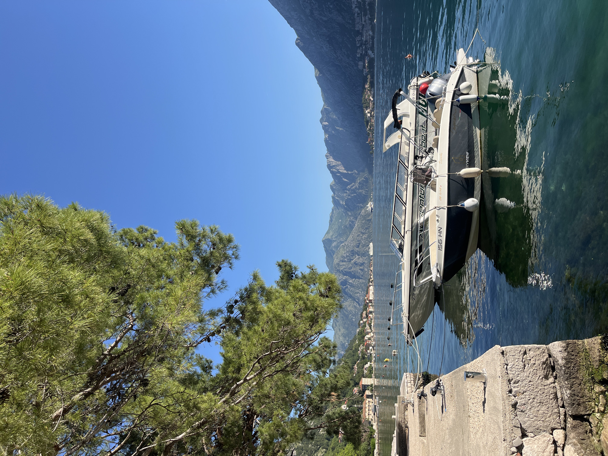 A boat is docked in the water next to a concrete pier. The boat is white and black. Mountains are in the background. The sky is clear and blue. A tree is to the left of the pier. The location is likely Škaljari, Montenegro, based on the architecture and environment visible.