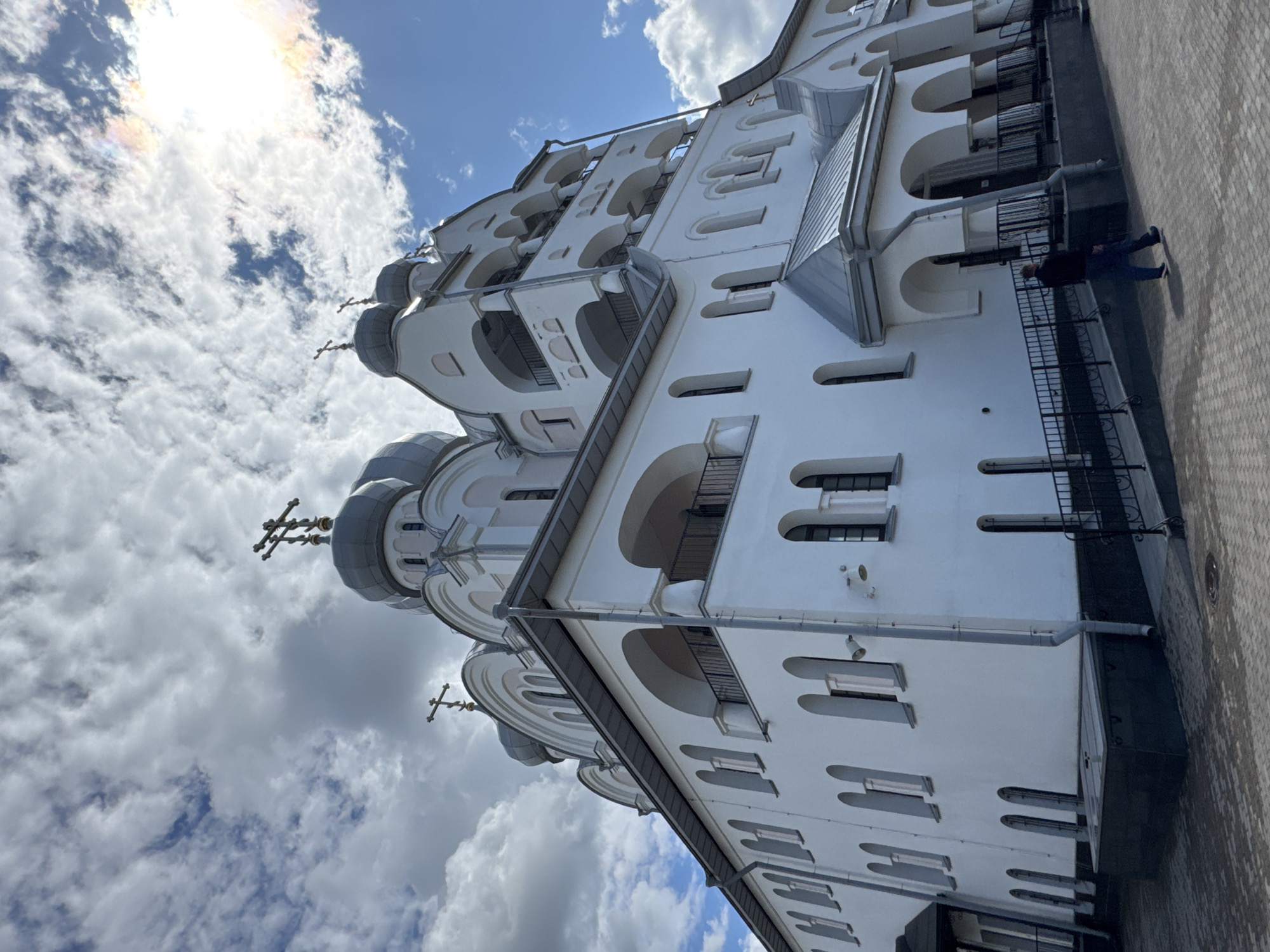 The image shows the Trinity Cathedral of the Izmaylovsky Regiment in Saint Petersburg, Russia, on a cloudy day. The cathedral is a large, white, multi-domed Orthodox church with multiple levels and arched windows. Several crosses are visible on the domes. The foreground shows a paved courtyard with a few security cameras and a ramp with a black metal railing. A single man, wearing dark pants and a dark jacket, is walking away from the viewer down the ramp towards the cathedral's entrance. The sky is partly cloudy with patches of blue sky and some sunlight breaking through, creating a slight rainbow effect in the upper right corner.