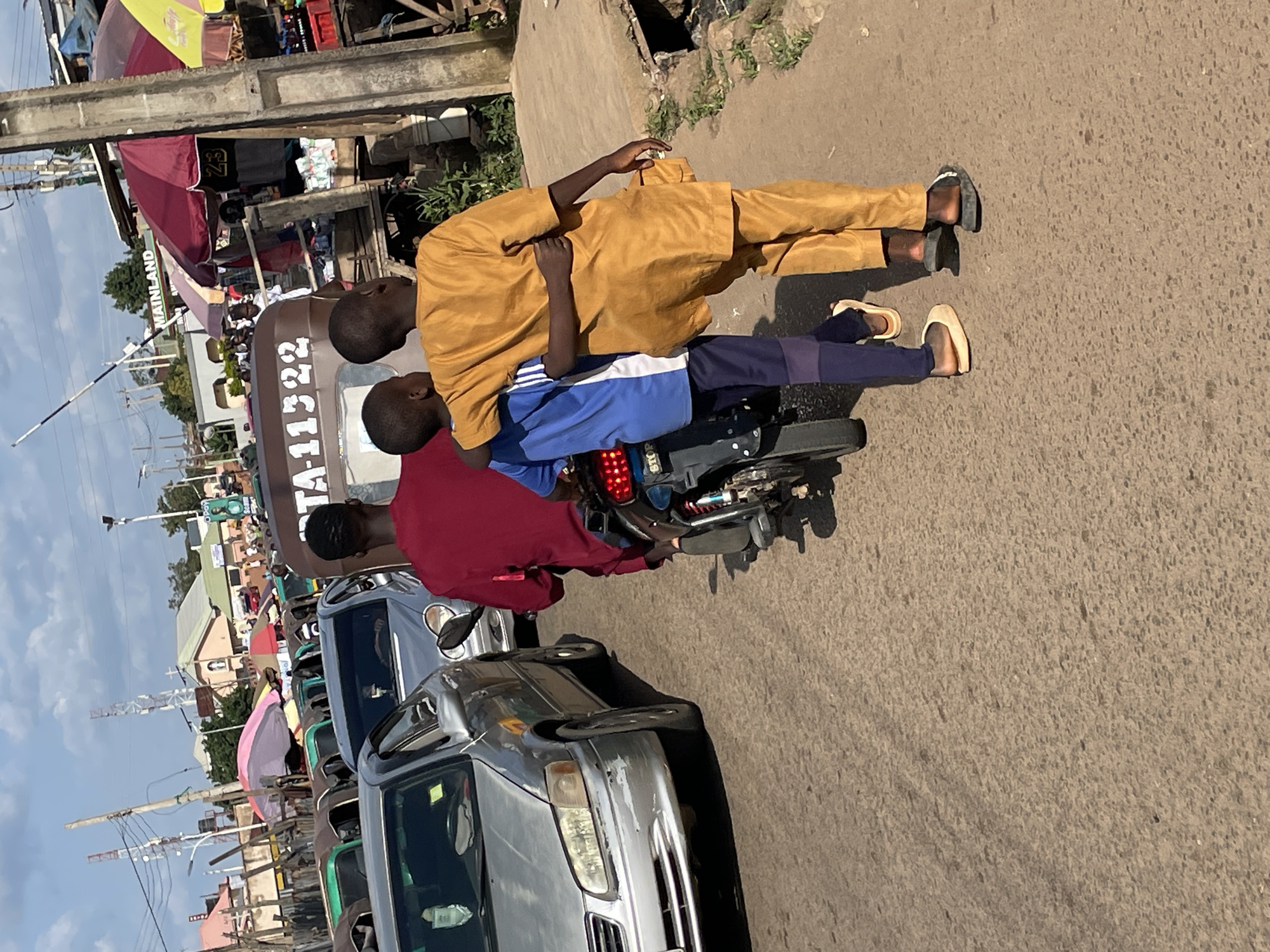 In Gura Top, Nigeria, two young boys are seen walking together with their arms around each other's shoulders on a crowded street. One boy is wearing a yellow traditional outfit, and the other is in a blue and white outfit. They are walking on the right side of the road, passing by a three-wheeled vehicle with the registration number TA-11322. The street is filled with various vehicles, including cars and the three-wheeled transport, suggesting a busy urban environment. On the right side of the road, market stalls with umbrellas and goods are visible, indicating a commercial area. The background shows buildings and communication towers, typical of a Nigerian city. The sky is clear with some clouds, and the sun is casting shadows, suggesting it is daytime. The overall scene depicts a vibrant, bustling street in Gura Top.