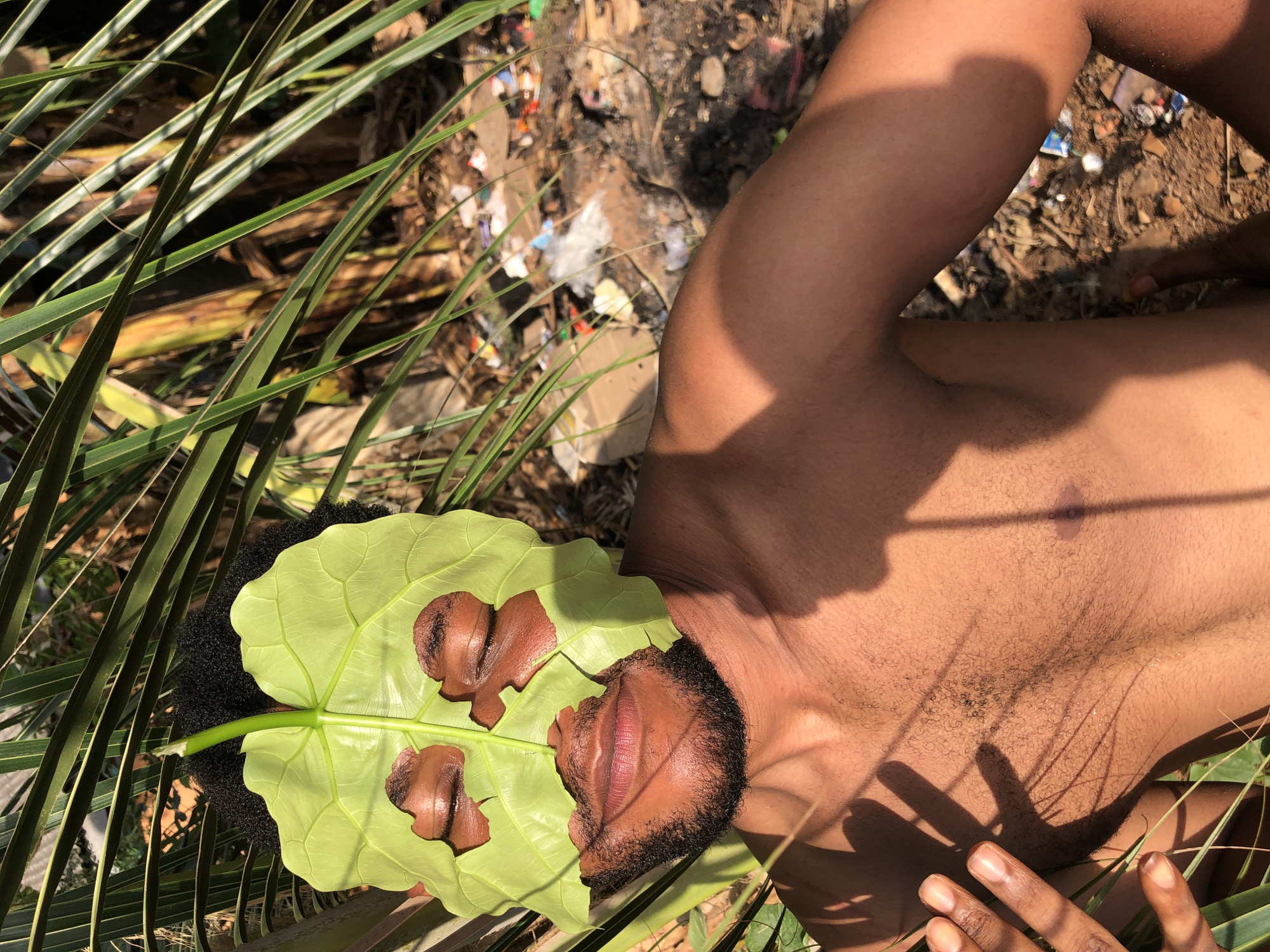 A high-angle, eye-level photograph shows a Black person lying on the ground covered with a large green leaf, palm fronds, and debris. The person’s head and torso are visible. A large, light green leaf covers the area of the person's eyes, and what appears to be their nose has been placed on top of the leaf. Only their mouth and beard are visible below the leaf. The person's dark hair is visible behind the leaf. The right arm is bent and visible to the right. A hand is also visible to the lower right. The person is lying among palm fronds. Beyond them, there is dirt, sticks, and debris. The location is listed as Unknown, Unknown.