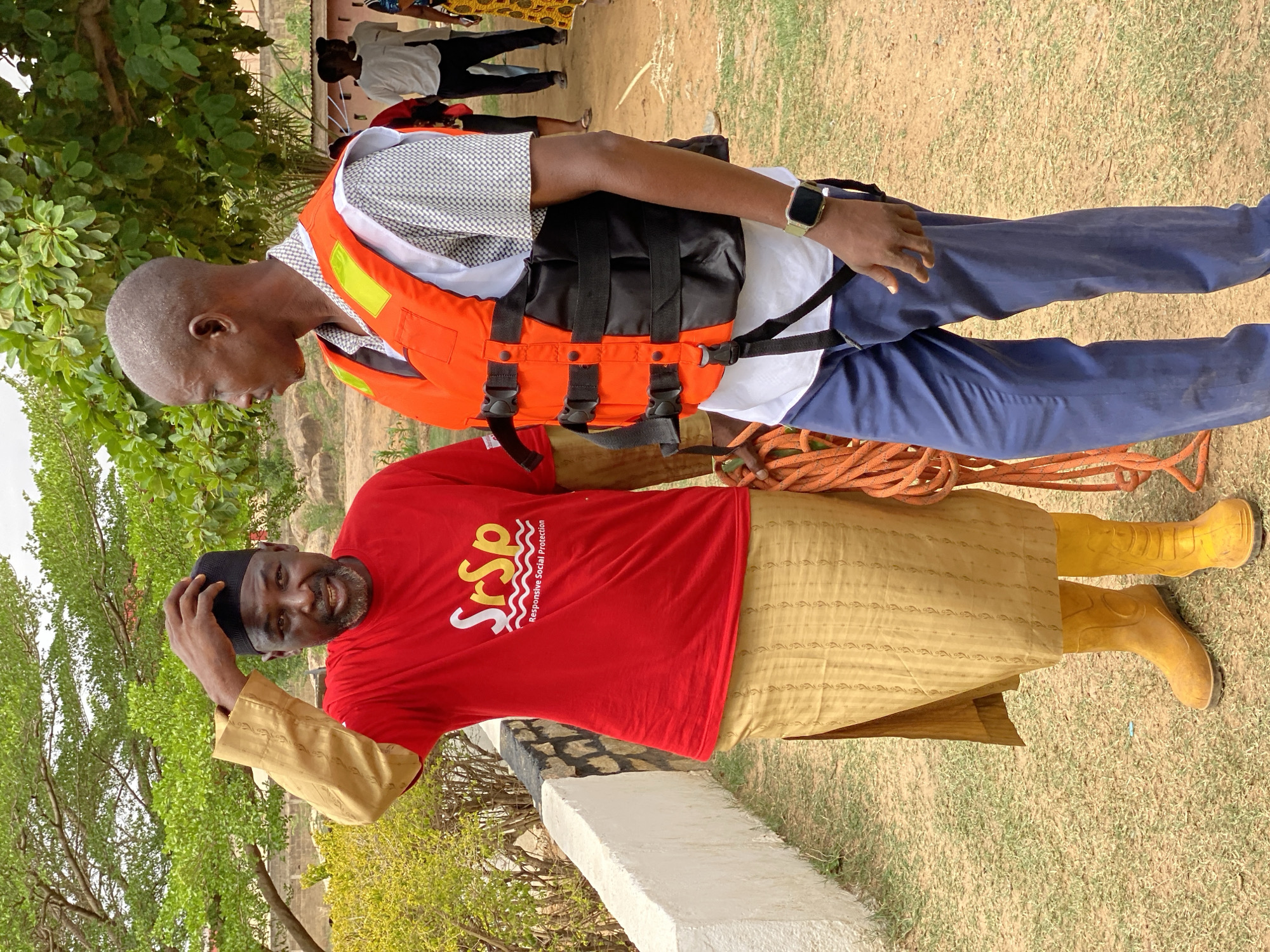 The image shows two men standing outdoors in what appears to be a grassy area with trees and other vegetation in the background. The location is in Tundun Wada South, Nigeria.
The man closer to the camera is wearing a red t-shirt with the acronym "SrSp" and the words "Responsive Social Protection," a light-brown robe-like garment, boots, and a dark-colored cap. He has a beard and is touching his cap with his right hand. He is smiling.
The other man is wearing an orange life vest over a white collared shirt and blue pants. He is looking down at the man in red. A rope is draped over the shoulder of the man in red. Other people are visible in the background but they are further away and less clear. The immediate surroundings consist of greenery and a low concrete structure. The ground appears to be a mix of grass and dirt.