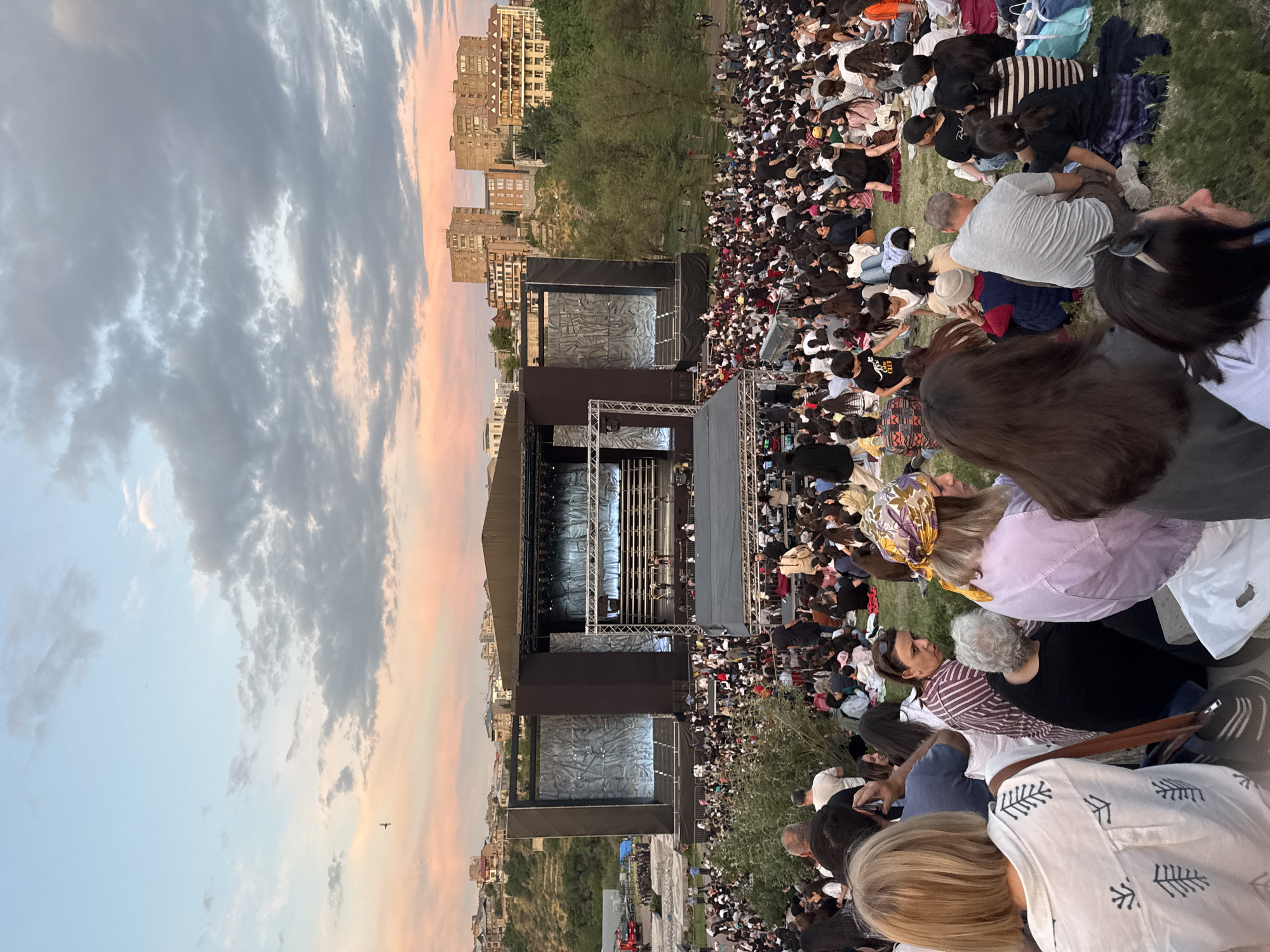 This image depicts a large outdoor gathering at dusk, focused on a concert or performance stage situated on a sloping, grassy hillside in Yerevan, Armenia. The sky transitions from a soft pink and orange sunset glow near the horizon to a lighter blue overhead, with scattered grey clouds. A single dark bird is visible in the upper left sky.
In the background, behind and to the sides of the stage, residential buildings are visible on elevated terrain. On the left, houses with red roofs and more uniform structures blend into a green hillside. On the right, larger, multi-story concrete residential buildings of typical post-Soviet architectural style stand against the sky, interspersed with trees. A small red utility or emergency vehicle is parked in the backstage area to the left of the main stage.
The central feature is a substantial, dark-colored outdoor stage structure. It comprises a lower, wider platform mostly obscured by the audience, and an elevated, smaller central performance platform where several indistinct figures (performers or crew) are present. The stage is flanked by two large, textured grey or white panels with undulating patterns, possibly serving as backdrops or screens. Metal trussing with lighting equipment is visible above and around the stage.
Hundreds of people are seated directly on the green grass of the hillside, facing the stage. They are densely packed, with individuals of varying ages visible, dressed in casual attire such as t-shirts, shirts, and jeans. Many have brought blankets or mats to sit on. The crowd extends widely across the visible grassy area, indicating a significant attendance. A large green bush partially obstructs the view of the lower stage and a section of the audience from the immediate foreground.
In the immediate foreground, several individuals are sharply focused, viewed from behind or in profile. On the far left, a person with blonde hair wears a white shirt featuring black tree-like graphic patterns. Next to them, further into the frame, a person with dark hair wears a striped shirt (reddish-brown and white) and looks towards the right. Close by, another person with grey hair is seen from behind, wearing a black top. On the right, a person with blonde hair wears a patterned headscarf (yellow, purple, green) and a light purple outer garment. The grass in the foreground shows wear from the presence of people.