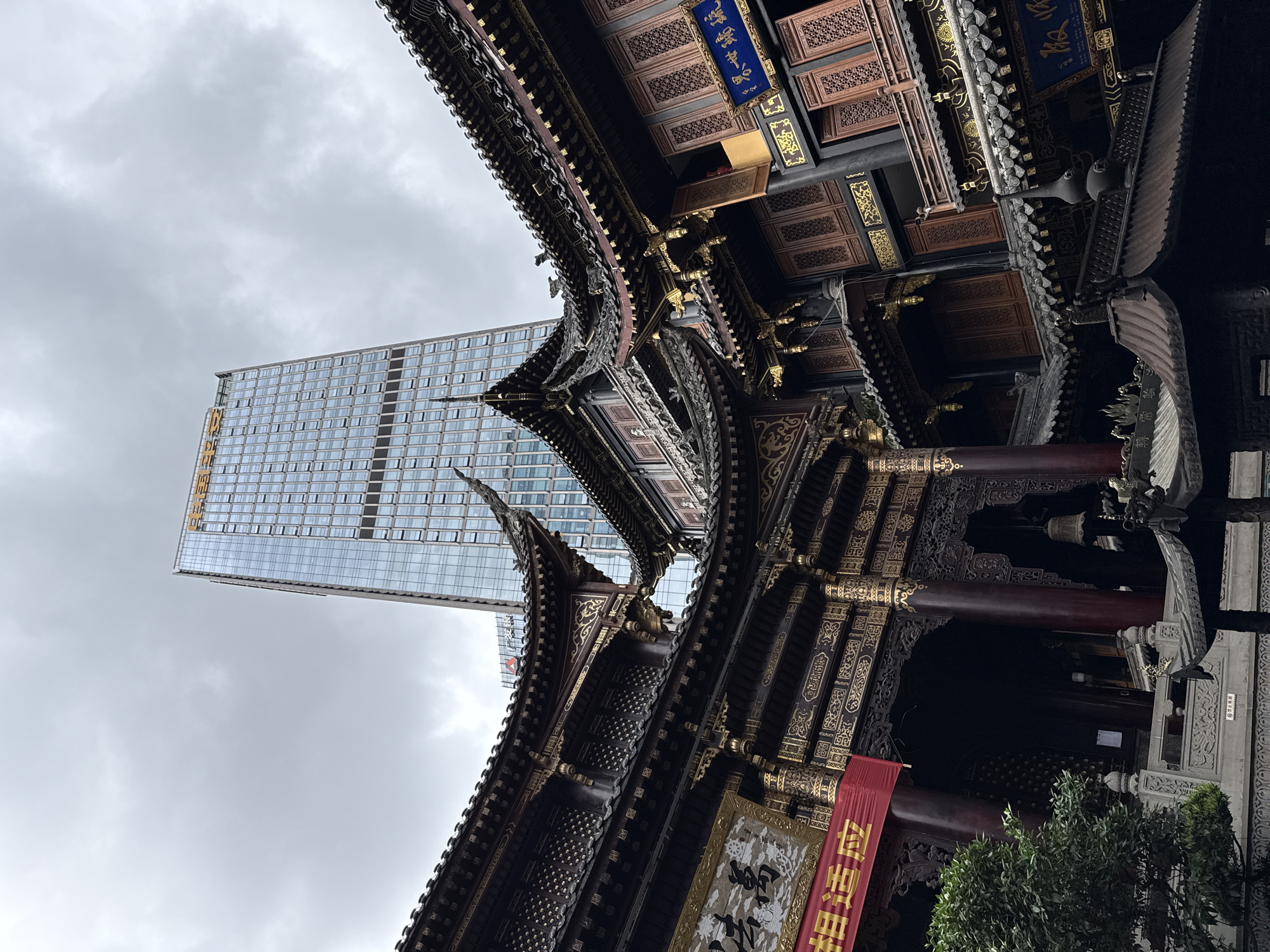 The image displays a striking architectural contrast under an overcast sky in Yuzhong District, China.
On the right side, a multi-tiered traditional Chinese building, likely a temple or pavilion, stands prominently. Its dark, ornate roofs are adorned with intricate carvings, golden accents, and decorative elements. The structure features rich wooden patterns, gold-painted trim, and dark red columns. A blue plaque near the top bears Chinese characters, appearing to read "重慶羅漢寺" (Chongqing Arhat Temple) and "大雄寶殿" (Mahavira Hall), indicating a Buddhist temple. A red banner hanging from a column in the lower right displays the characters "佛光普照" (Buddha's light shines universally).
To the left and behind this ancient edifice, a sleek, modern glass skyscraper soars into the gray sky. Its facade is a grid of reflective windows, and a subtle yellow logo or text is visible vertically on its upper-left side.
The sky above is entirely covered by soft, diffuse gray clouds, suggesting an overcast daytime. No people or specific activities are visible, emphasizing the architectural subjects. The photograph effectively juxtaposes China's historical heritage with its contemporary urban development.