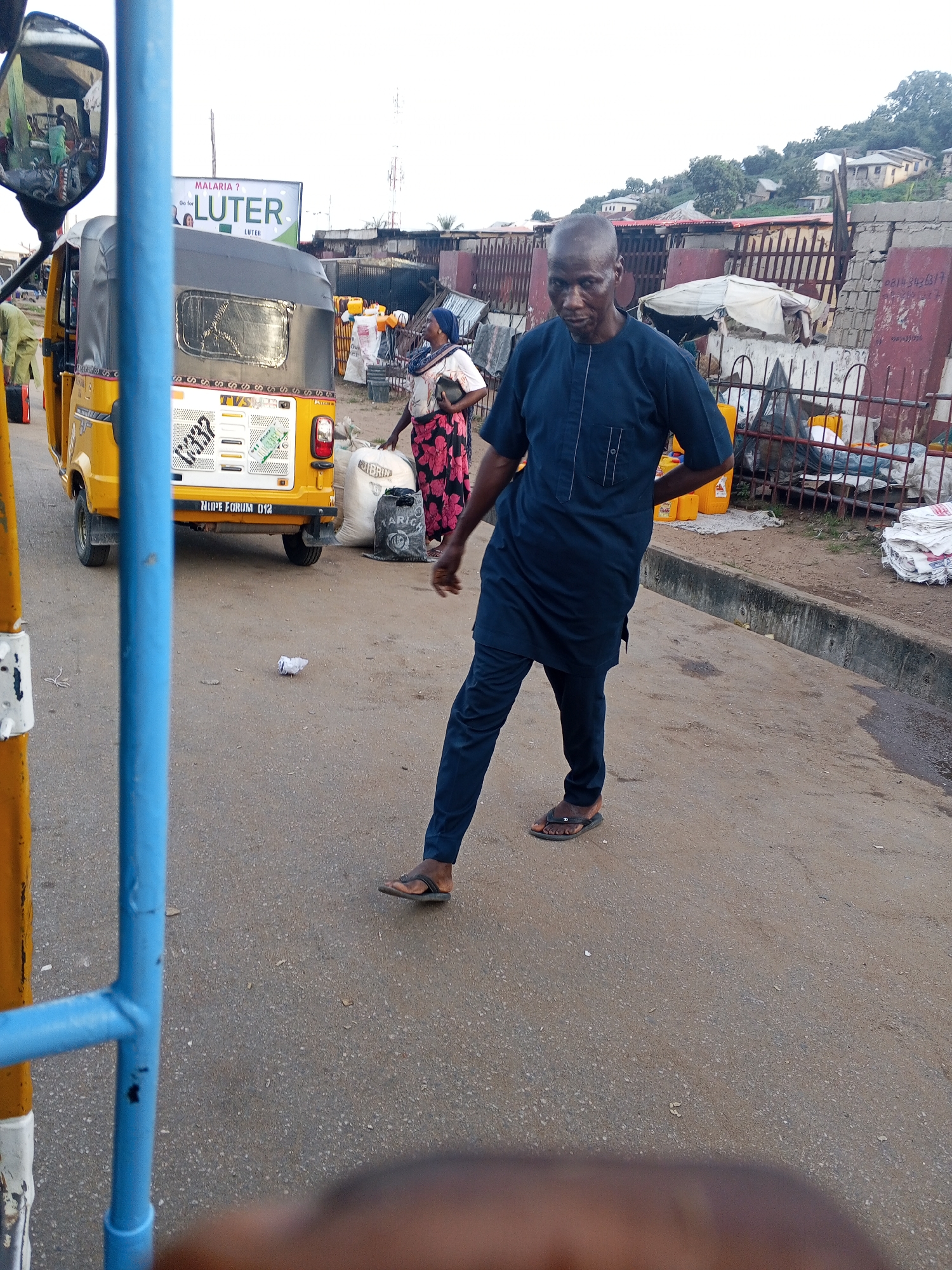 A man wearing a dark blue traditional outfit is walking on a paved street in Lokoja, Nigeria. He is facing right and appears to be looking towards the right. To his left, a yellow auto-rickshaw is parked. Behind the auto-rickshaw, a woman in a black and floral patterned dress is standing near some sacks. In the background, there are buildings and a billboard that reads "MALARIA? Or for LUTER LUTER". The sky is overcast, and the scene appears to be a street market or a busy roadside area.