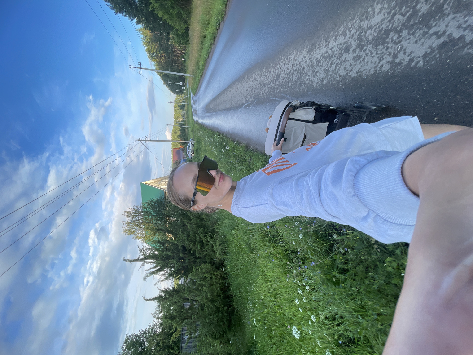A person is taking a selfie while walking a stroller on a wet, rural road in the village of Podshivalovo, Russia. The person, wearing large sunglasses and a light-colored sweatshirt with orange lettering, is positioned in the foreground, with their arm extended to capture the shot. To their left is a grassy area with wildflowers and several conifer trees, and a house with a green roof is visible in the background. To the right, the road curves into the distance, flanked by trees and power lines. The sky is partly cloudy with a blue hue. The stroller, a light-colored model with a dark frame, is positioned beside the person.