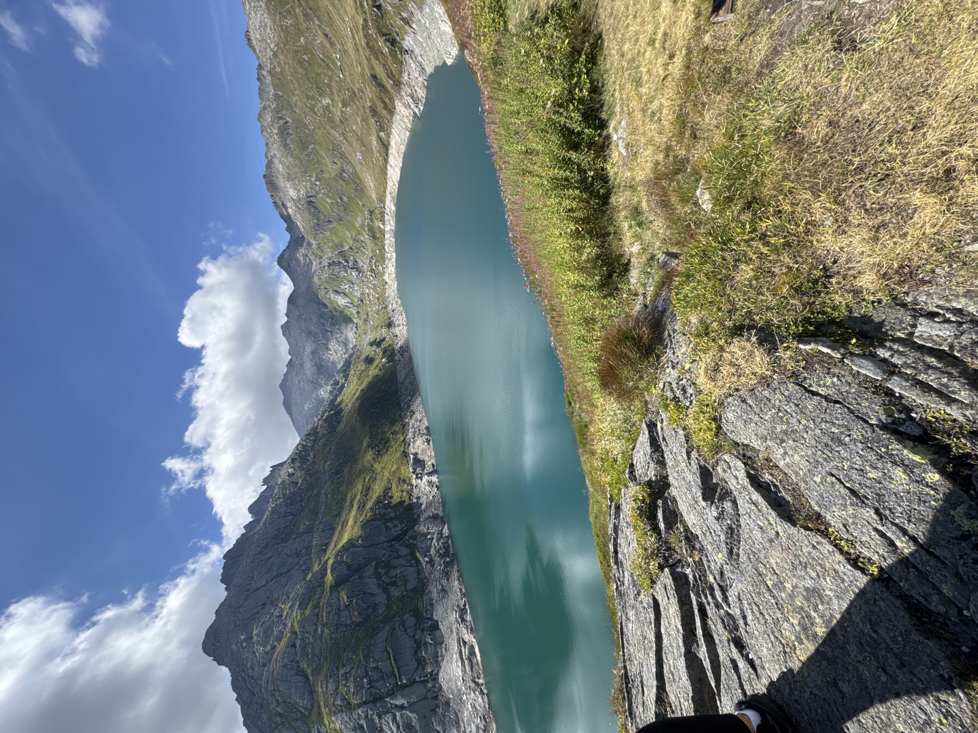 The image shows a scenic view of a turquoise mountain lake surrounded by rocky, green slopes under a bright blue sky with scattered clouds. In the foreground, large, weathered grey rocks are visible, with sparse vegetation growing between them. The lake itself is calm, reflecting the sky and surrounding mountains. The background features rugged mountain peaks, some bare rock and others covered in green grass. The scene is indicative of a high-altitude natural environment. The location is identified as the Lago di Luzzone in the municipality of Airolo, Switzerland.