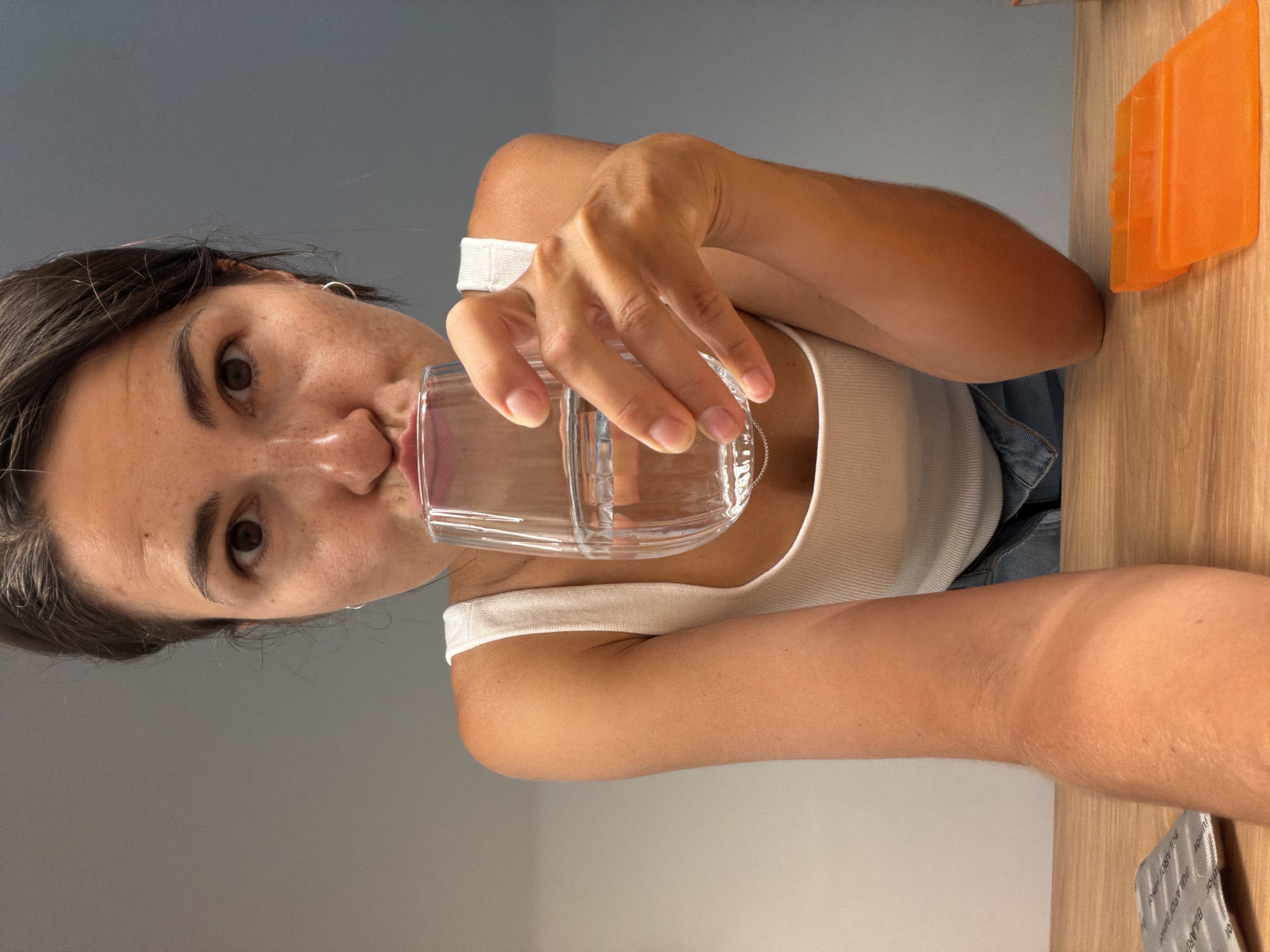 A woman is holding a glass of water and about to drink it. She is wearing a white tank top and denim shorts. In the foreground, there is a pill packet and an orange object on a wooden surface. The background is a gray wall. There is no information available about the location being Budva, Montenegro.