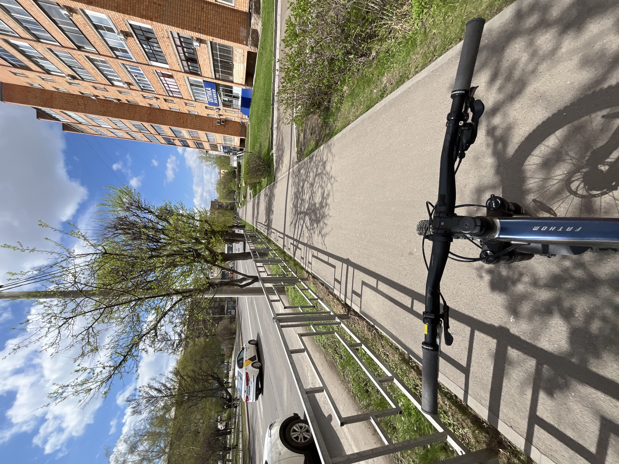A first-person perspective image of a bicycle ride along a sidewalk in Krasnoyarsk, Russia. The handlebars of the blue "Fathom" bicycle are in the foreground. The sidewalk runs parallel to a road with parked cars, separated by a metal fence. On the other side of the sidewalk, there is a brick apartment building with a blue sign written in Russian on the side. The sky is blue with scattered clouds. Trees line the sidewalk and road. The shadow of the bicycle's front wheel is cast on the sidewalk.