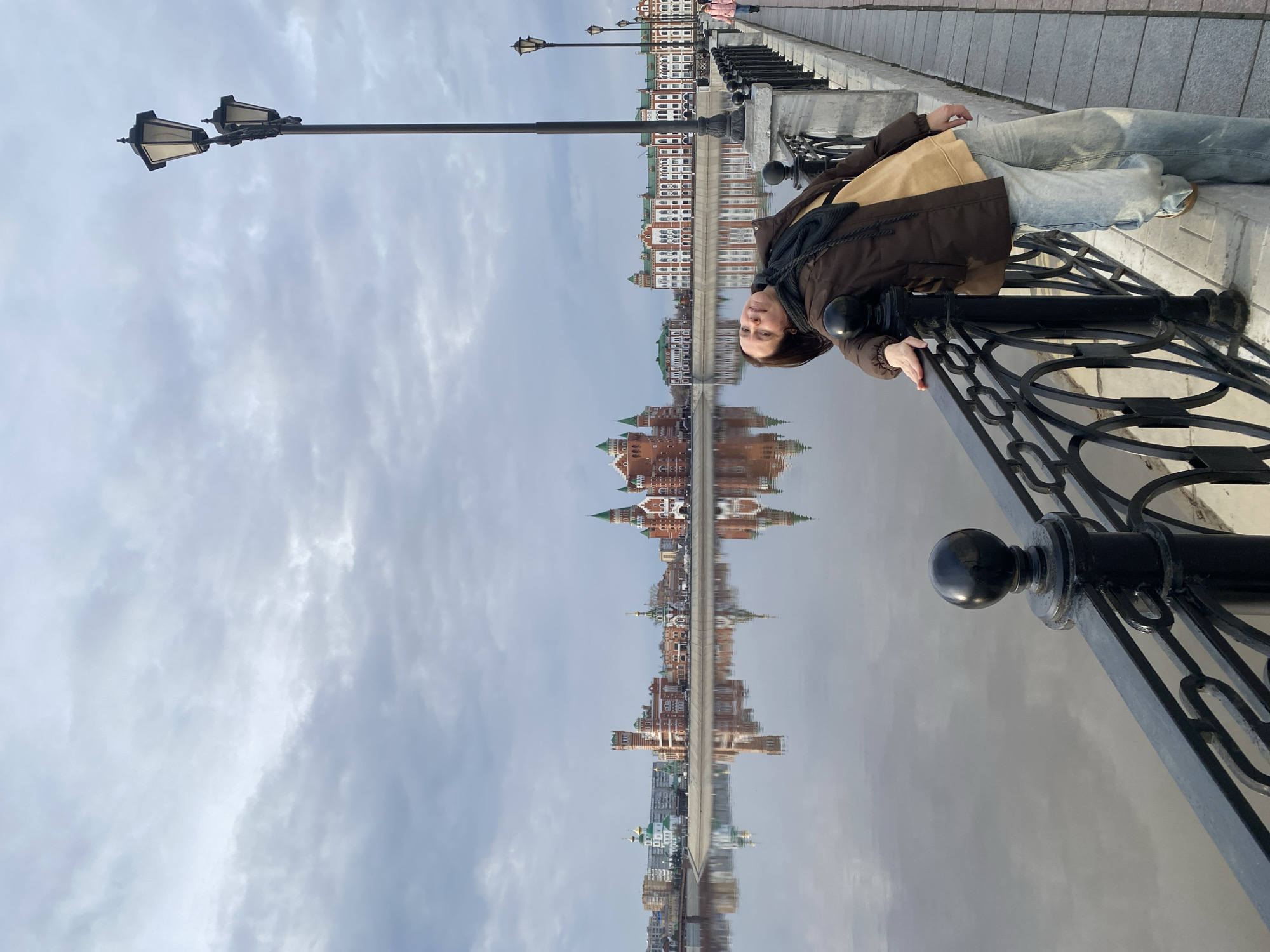This full-length, eye-level photograph captures a serene city embankment scene featuring a person in the foreground. A woman, dressed in a brown jacket, light blue jeans, and a dark scarf, is positioned on the right side of the frame. She is casually leaning against a black ornate metal railing, with her hands resting on it, and looks directly towards the camera with a neutral expression.
The railing extends diagonally from the bottom right, separating the paved walkway from a calm body of water, likely a river or canal. The water perfectly reflects the cityscape on the opposite bank, creating a striking mirrored effect. Visible across the water are distinct buildings, characterized by red brick facades and prominent green roofs, consistent with the architectural style found in Yoshkar-Ola, Russia. Their reflections are clear and vivid in the tranquil water.
Above the cityscape and its reflection, a soft, overcast sky with diffused clouds fills the upper portion of the image. Street lamps line the paved embankment behind the woman, receding into the distance. The lighting suggests a cloudy daytime, creating a soft and even illumination. There is no visible text in the image.