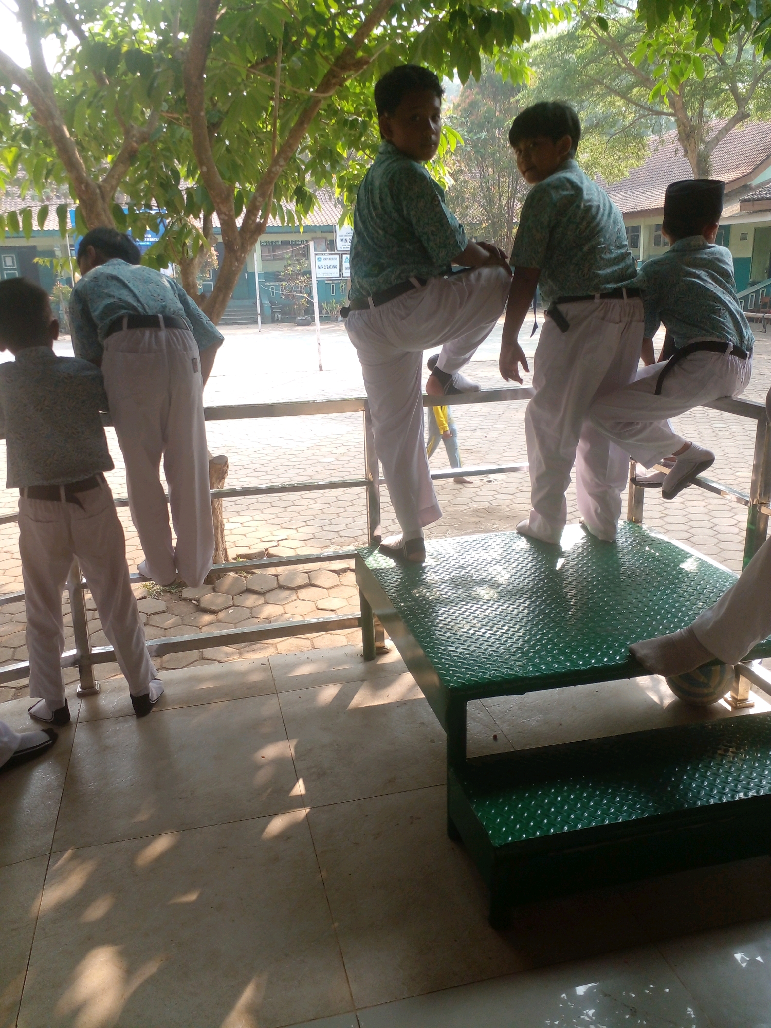 The image depicts five young boys, likely students in school uniforms, on an outdoor paved area with trees and buildings in the background. The boys are wearing light-colored shirts with a patterned design and white pants. Some are standing, some are sitting on a green metal platform with a textured surface. One boy on the far right is partially visible, wearing a black peci or skullcap. The environment appears to be a schoolyard, possibly in Banyuputih, Indonesia, given the presence of buildings and mature trees casting shadows on the ground. Sunlight filters through the leaves, creating dappled patterns on the pavement. A soccer ball is visible near the green platform. The overall scene suggests a casual moment during a school day.
