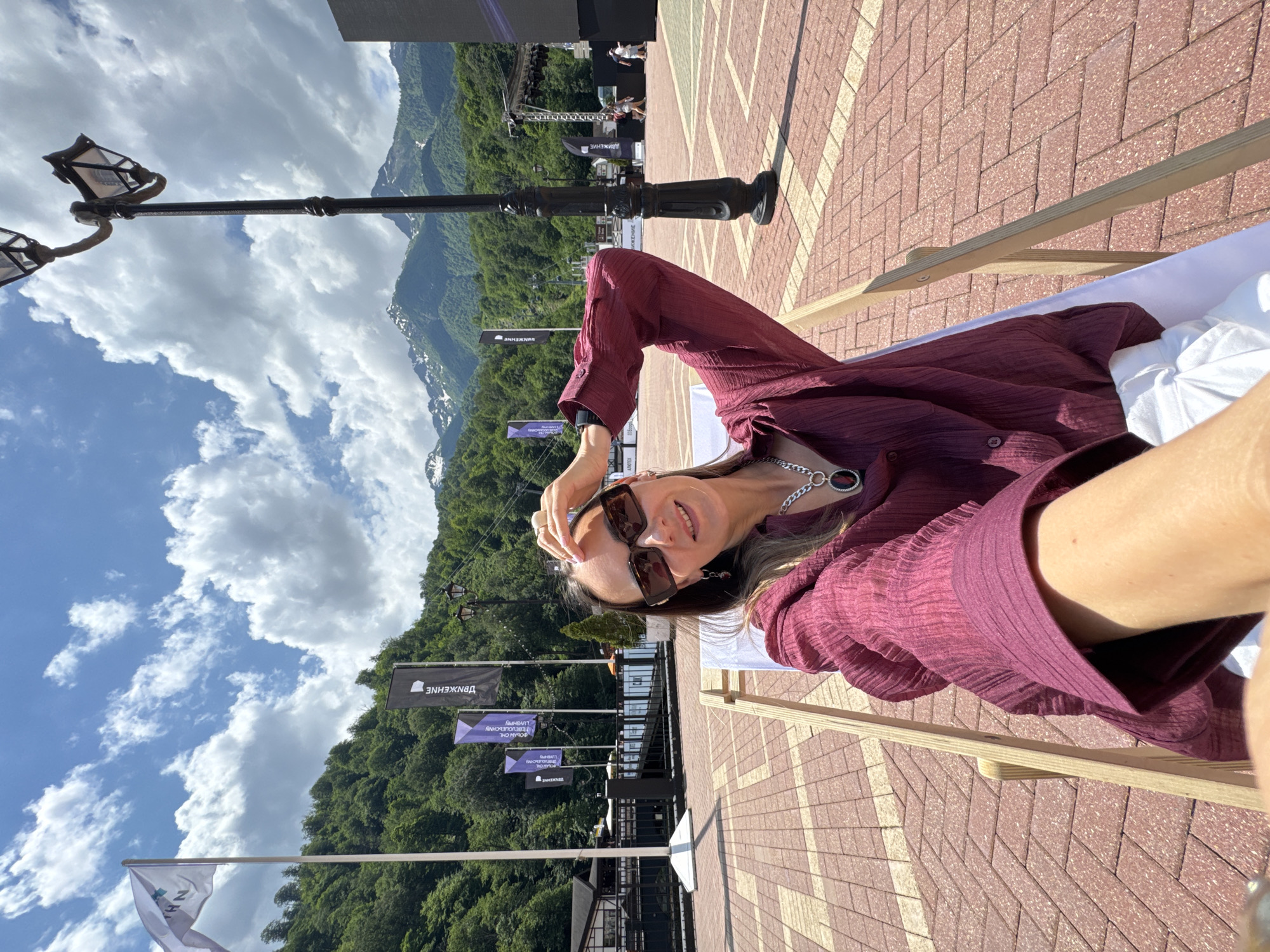 A woman is taking a selfie while sitting in a deck chair outdoors. She is wearing sunglasses, a burgundy-colored shirt, and white pants. She has her right hand on her head. The background shows a paved area with red and brown bricks, flags, a lamppost, trees, and snow-capped mountains. The scene is set in Estosadok, Russia.