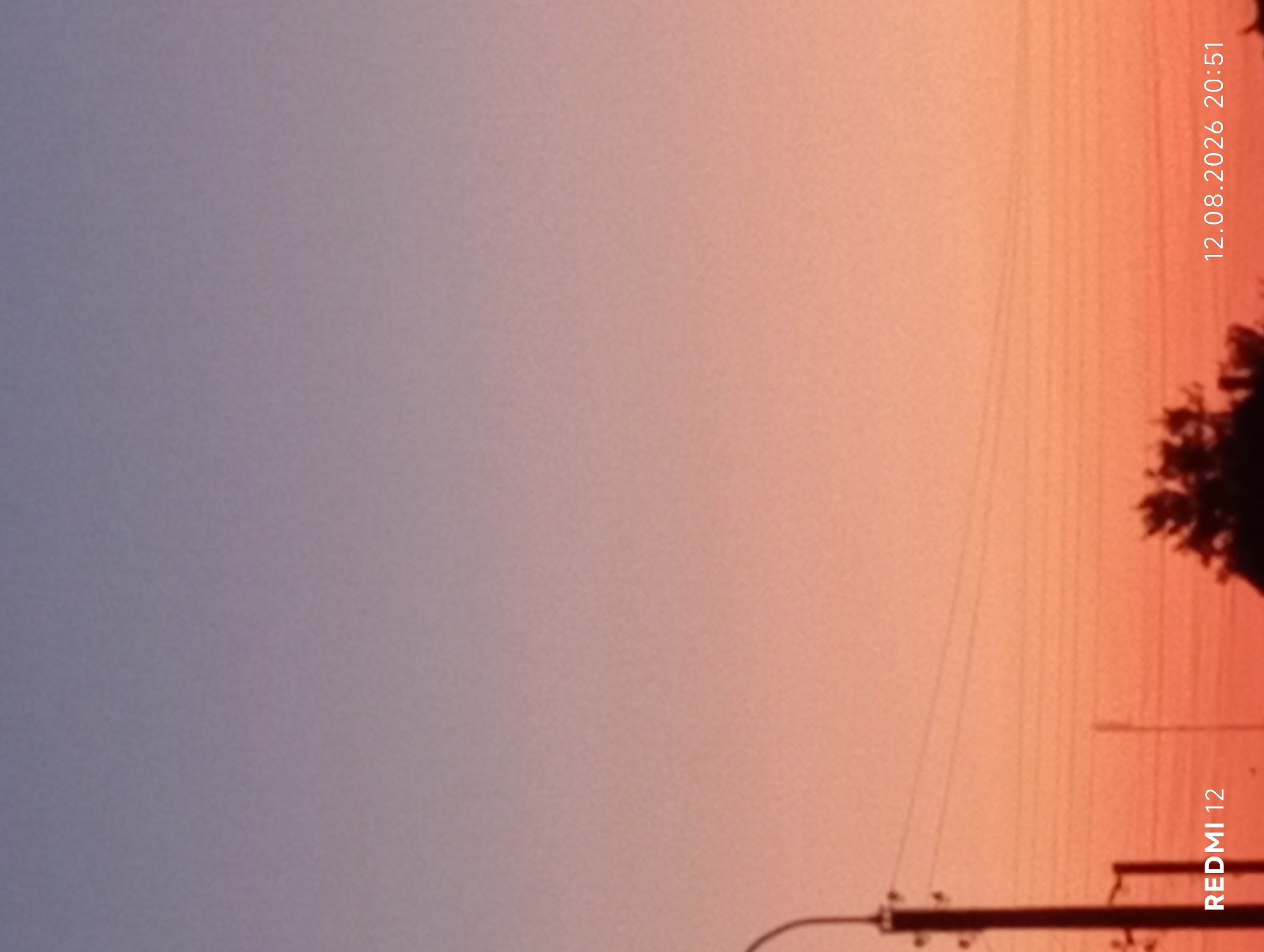 This is a photograph taken in the city of Smila, Ukraine. The image captures a twilight sky with a gradient of colors from purple and blue at the top to orange and pink near the horizon. Silhouetted against the vibrant sky are power lines, a street lamp pole, and the tops of trees. The bottom left corner of the image displays the text "REDMI 12", indicating the device used for capture. The bottom right corner shows the date and time: "12.08.2026 20:51". No people are visible in the frame.