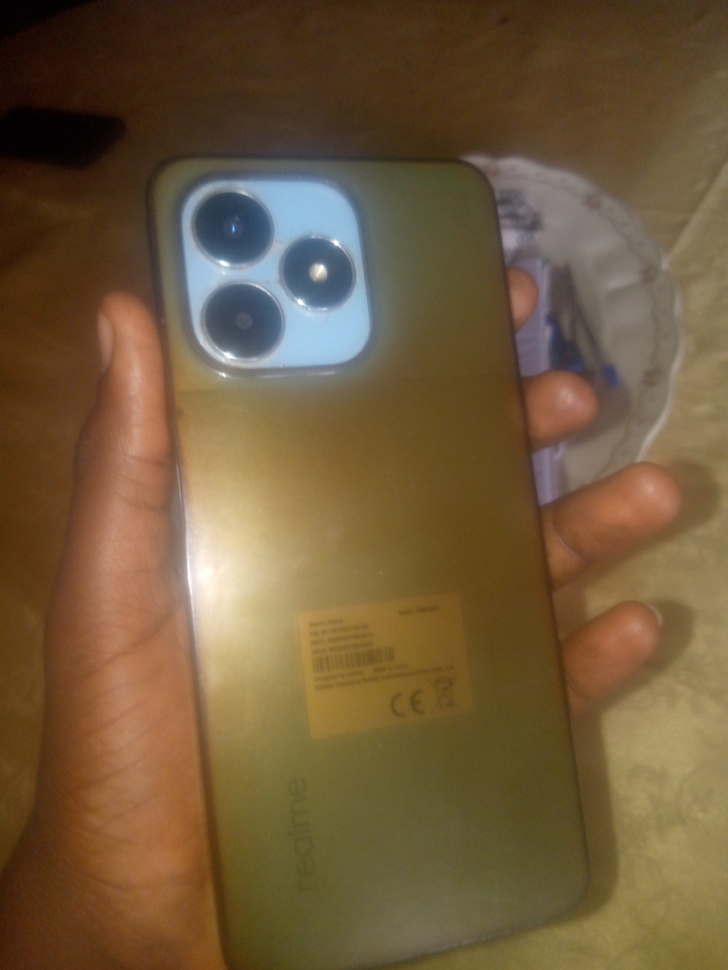A person's hand holds a gold-colored smartphone. The smartphone has a triple-lens camera system on the back. A label with "Realme" logo is visible on the back of the phone. The phone is held in a person's hand, covering the lower right corner of the image. The background is a patterned surface, possibly a bedspread or tablecloth, in shades of yellow and brown. The lighting is dim, casting shadows. There is no specific information to determine the location in the city of Jauro Yinu, Nigeria, from this image alone.