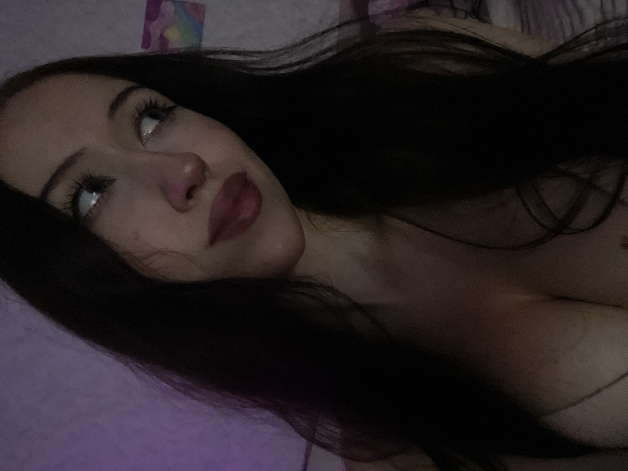 This is a low-angle, close-up, portrait-style image of a young person with long, dark hair. The individual's face is turned upwards and slightly to the left, with their eyes looking in the same direction. They are wearing dark makeup, including mascara and lipstick. Their hair is dark and appears to be long, framing their face and extending out of frame.
The background is dimly lit and appears to be indoors. There are hints of purple lighting in the lower left corner and a small, colorful, abstract image is visible on the wall in the upper left. The scene suggests a casual or intimate setting, possibly a bedroom. The overall mood is contemplative or dreamy, enhanced by the soft lighting and the subject's gaze. There is no visible text in the image. The time of day is likely evening or night, given the dim lighting.