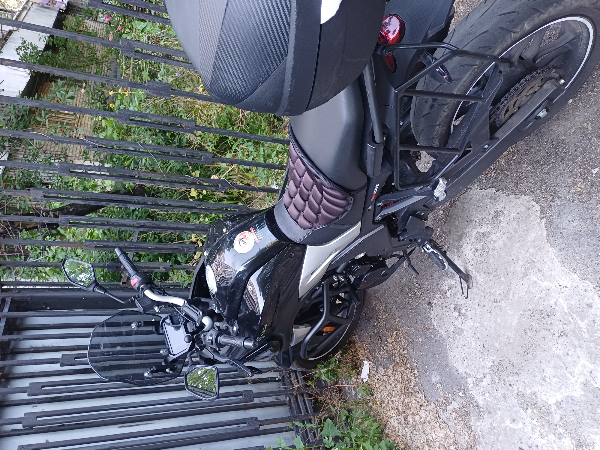 A black motorcycle is parked next to a black metal fence. The motorcycle has a black seat with a diamond-patterned insert, a black top box with a textured surface, and a black windscreen. It also has a black engine guard. The motorcycle is parked on a paved surface with some gravel and greenery visible. The scene appears to be outdoors during the daytime. The fence is partially covered by green foliage, suggesting a natural or semi-natural environment. The overall impression is that of a parked vehicle in an urban or suburban setting. No people are visible in the image. The image is taken from a slightly elevated angle, looking down at the motorcycle.