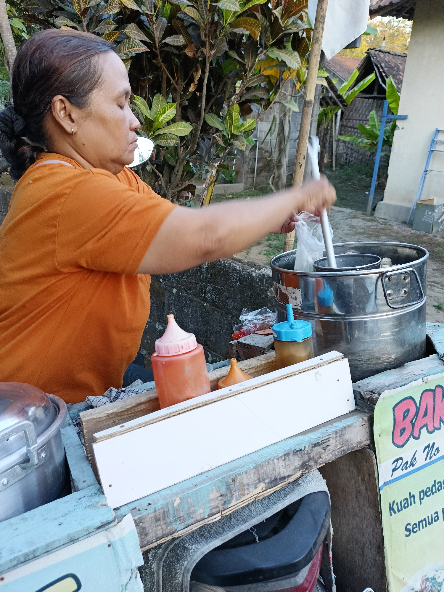 A medium shot captures a woman in an orange t-shirt with her eyes closed, preparing food at a street stall in Borangan, Indonesia. Her hair is pulled back into a bun, and she wears a small hoop earring. She is using a metal ladle to scoop something from a large stainless steel pot into a plastic bag. Beside the pot are several condiment bottles, one orange and two smaller ones with blue and brown caps. The stall itself is made of worn, painted wood, with a white board in front. Behind the stall, a stone wall and lush green foliage are visible, along with some traditional Indonesian houses and a blue ladder. A sign on the right side of the stall displays the words "BAK" in large pink letters, followed by smaller text. The lighting suggests it is daytime, possibly late afternoon, given the warm tones.