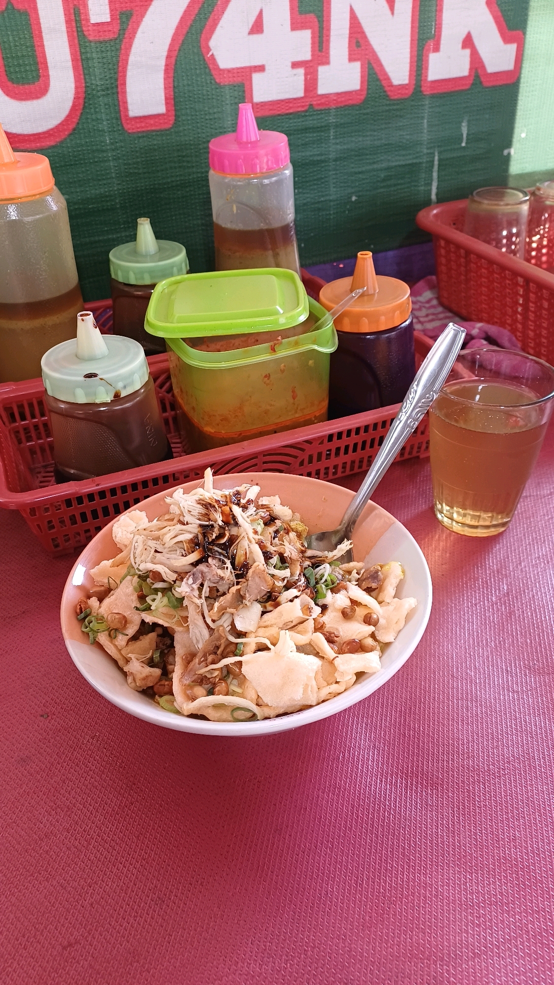 A close-up shot features a bowl of savory food, likely a local Indonesian dish, placed on a pink textured tablecloth. The dish is topped with shredded chicken, crispy crackers, and garnished with chopped green onions. A spoon rests in the bowl. To the left of the bowl are various sauce bottles, some with colorful caps, and a green plastic container with a lid, all arranged in a red basket. To the right, a clear glass contains a beverage, possibly tea. In the background, a green banner with large, red-outlined white lettering is visible. This scene suggests a food stall or a casual dining setting, potentially in Citeureup, Indonesia, a region known for its vibrant street food culture.