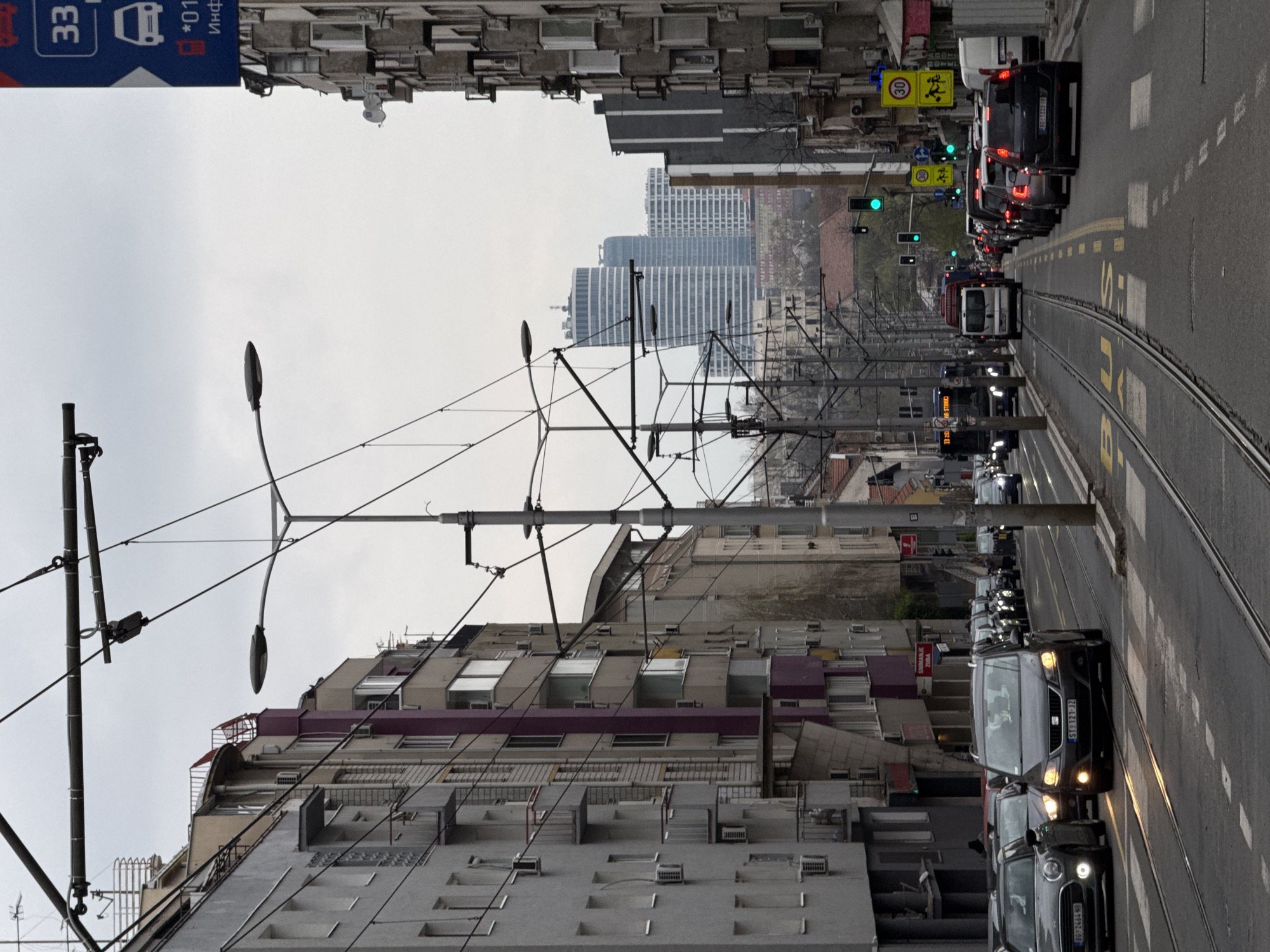 The image depicts a street in Vozdovac Urban Municipality, Serbia. The view is along a street with tram tracks running down the center. Traffic is present in both directions, with a line of cars on the right side of the frame. On the left, buildings line the street, interspersed with streetlights and tram power lines. In the distance, modern high-rise buildings are visible against a gray sky. The weather appears overcast.