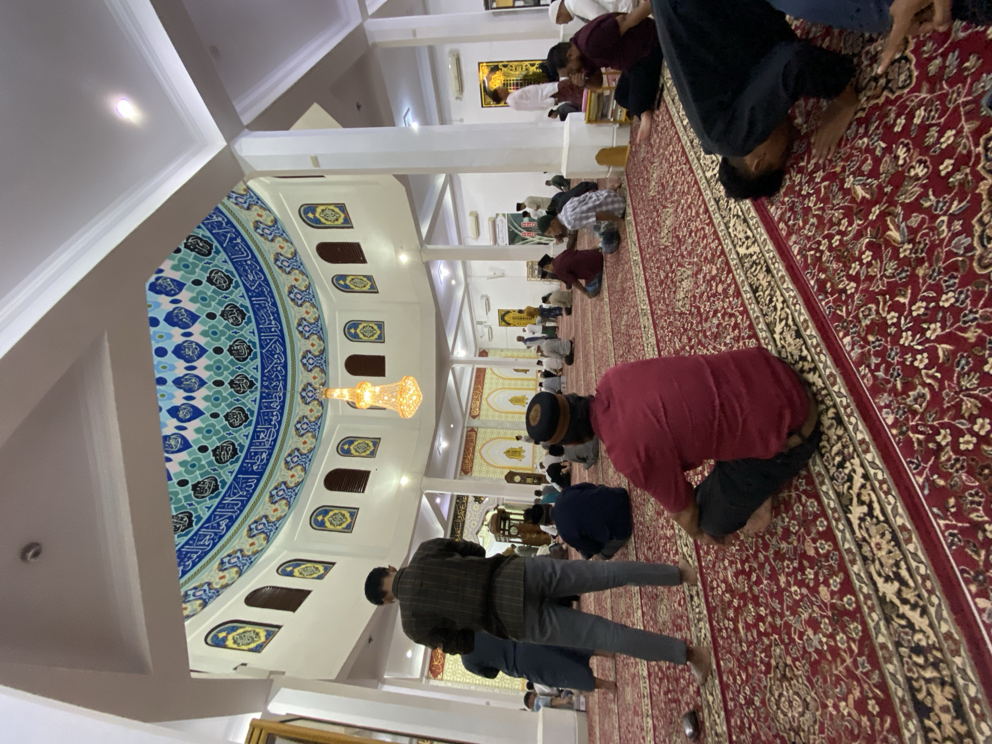 The image captures an interior view of a mosque in Lhokseumawe, Indonesia, during a prayer service. Numerous individuals are seated and kneeling on a large, intricately patterned red carpet. The architecture is adorned with decorative elements, including a domed ceiling featuring blue and teal geometric designs and Arabic calligraphy. Several arched windows are visible along the walls, some with decorative panels. A chandelier hangs from the ceiling, and pillars support the structure. People are dressed in casual attire, with some wearing traditional Islamic caps. The atmosphere appears solemn and communal, with individuals engaged in prayer.