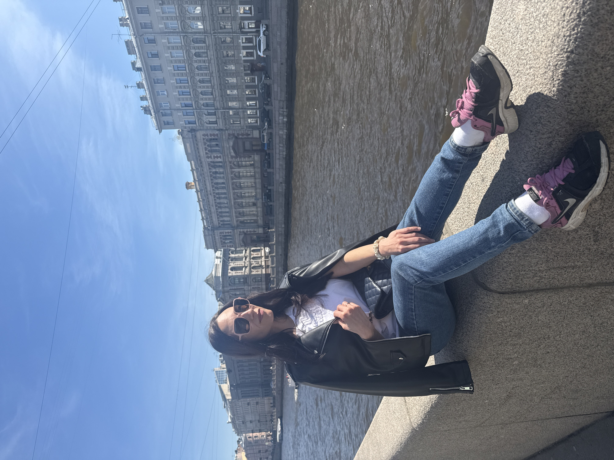 A woman is sitting on a stone embankment in Saint Petersburg, Russia. The location is likely near the river Neva given the buildings visible in the background.
The woman is wearing blue jeans, a white t-shirt, a black leather jacket, and black and pink sneakers with white socks. She is also wearing sunglasses. She is sitting with her legs extended in front of her, resting on the embankment.
Behind her is the river and across the river are buildings typical of Saint Petersburg architecture. The sky is clear and blue. The photo appears to be taken during the day.