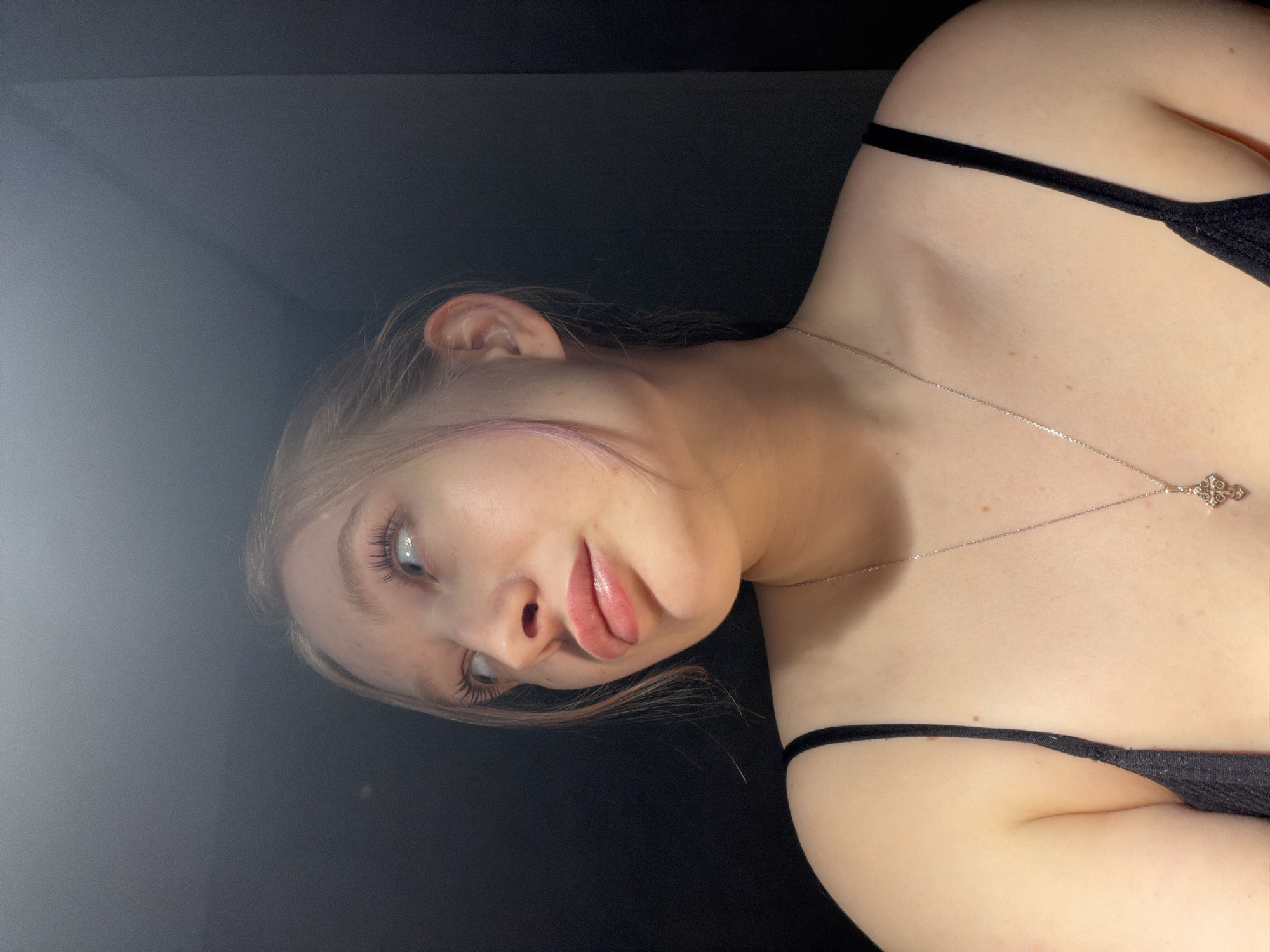 A pale-skinned young woman with light hair and blue eyes is depicted in a close-up, off-kilter perspective shot. She wears a black spaghetti-strap top and a silver necklace with a cross pendant. The background is a dark, nondescript setting with a possible corner in the upper left. The subject's expression is playful, with slightly pursed lips and eyes directed to the left. Location: Rossiyskiy, Russia is unknown from the image content.
