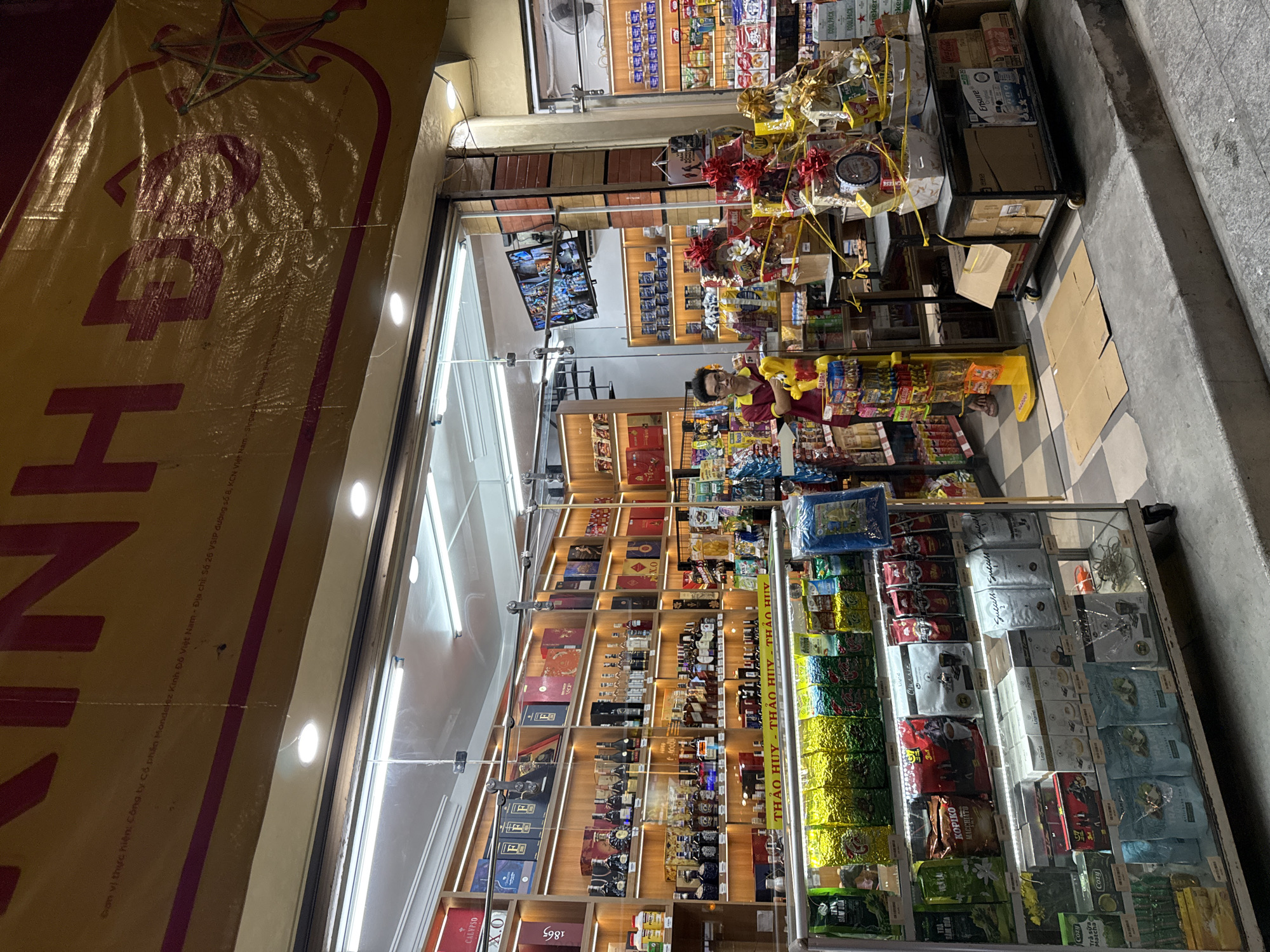 This is a medium shot of a convenience store in Nha Trang, Vietnam, at night. The store entrance is open, revealing shelves stocked with various goods, including bottles of alcohol, packaged food items, and gift baskets. A young male employee, wearing a red and yellow uniform, stands near the entrance, facing towards the street. A television screen mounted above the entrance displays a CCTV feed. In front of the store, several gift baskets are displayed on a cart, and a few cardboard boxes are placed on the sidewalk. The street in front of the store is paved with concrete. The overall scene suggests a busy commercial area.