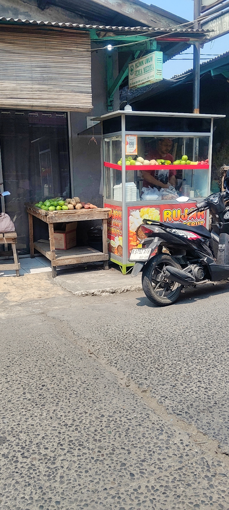 The image shows a street scene in Mekarmukti, Indonesia. In the foreground, a textured asphalt road is visible. To the right, a black motorcycle is parked. Behind the motorcycle is a small food stall, constructed with glass panels and a metal frame, featuring a red trim and signage that reads "RUJAK." The stall is stocked with various fruits, including green apples and what appear to be pears, and a stack of white plastic containers. A person, likely the vendor, is partially visible inside the stall, wearing a red shirt. To the left of the food stall is a wooden table laden with a variety of fresh produce, including green fruits, root vegetables, and sweet potatoes. Behind the table, a doorway is covered by a bamboo blind. Above the stall, a green sign with white text announces "KLINIK UMUM VEMIA MEDIKA" and provides operating hours. The overall environment suggests a typical street-side business in a residential or commercial area.