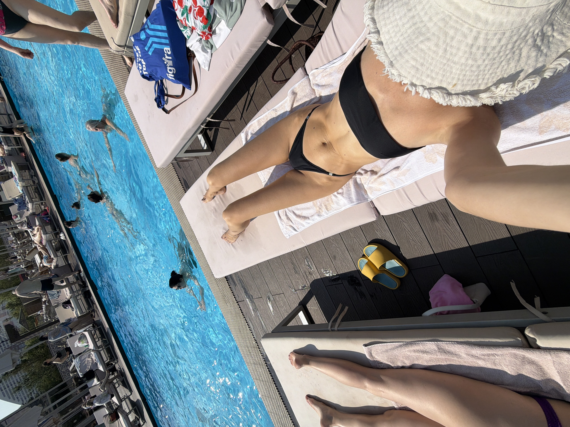 The image shows a sunny day at a pool, likely in Khimki, Russia, with people swimming and relaxing. In the foreground, two individuals are lounging on poolside chairs. One person is lying on their back on a pink towel, wearing a black bikini, and has a white fringed cover draped over their lower body. Their legs and torso are visible. Next to them, another person's legs are extended on a beige towel on a separate chair. A pair of yellow slides are on the wooden deck nearby, along with a pink cloth bag peeking out from under a chair. In the middle ground, several people are swimming in the bright blue pool. One woman with dark hair and sunglasses is swimming towards the foreground, while another woman with blonde hair is doing a backstroke further out. In the background, more people are visible around the pool, with lounge chairs and umbrellas. The overall environment suggests a recreational area, possibly a hotel or public pool.