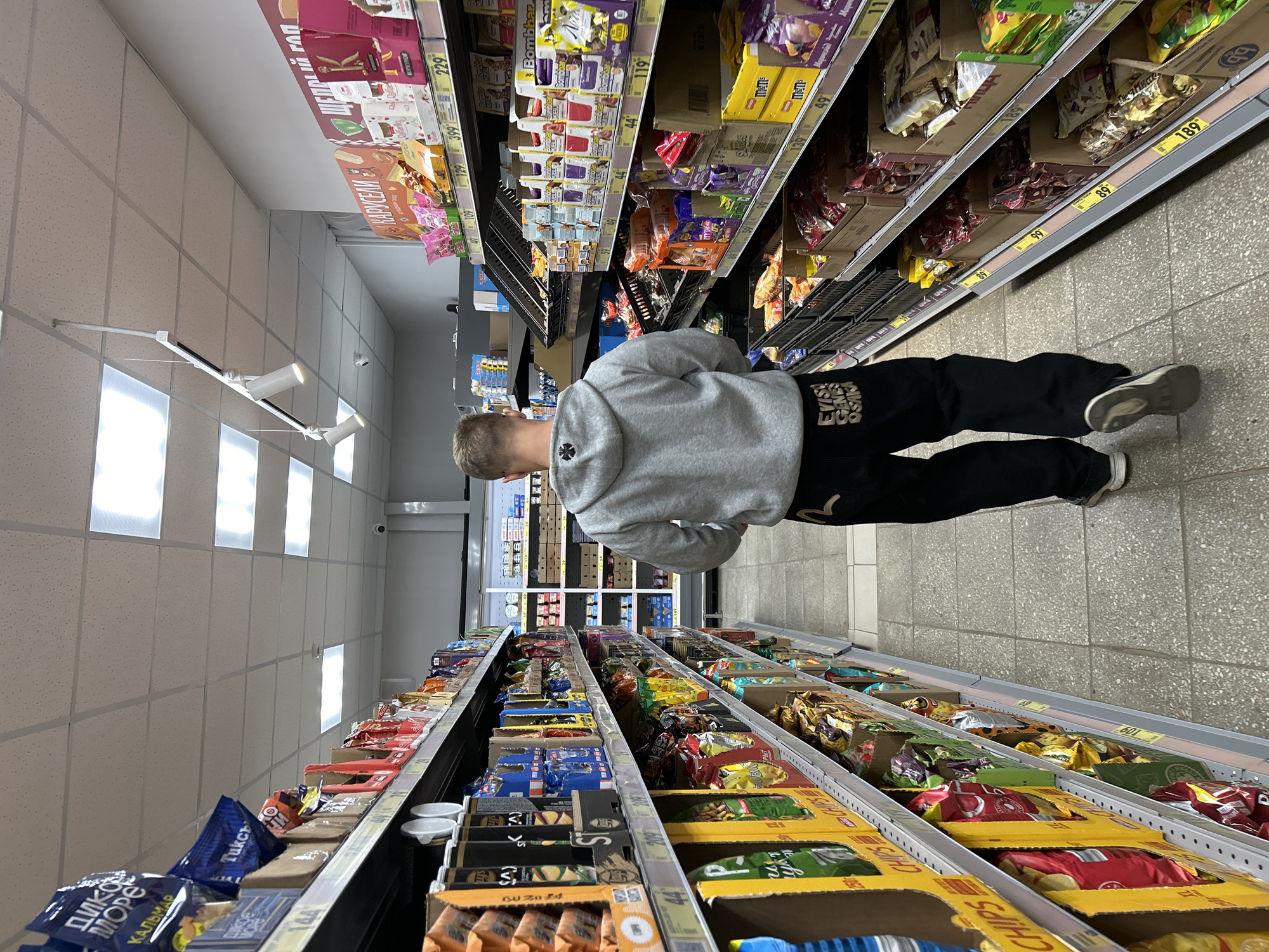 A young male is walking away from the camera down an aisle in a brightly lit, modern supermarket. He is wearing a grey hooded sweatshirt and black pants with an embroidered design on the back of the legs. The aisle is lined with shelves stocked with various snack food products. The shelves are organized and well-maintained, with price tags visible on some of the products. The floor is tiled with a speckled grey pattern. The overall impression is that of a typical grocery store shopping experience. The specific location in Moscow, Russia is not discernible from the visual information provided.