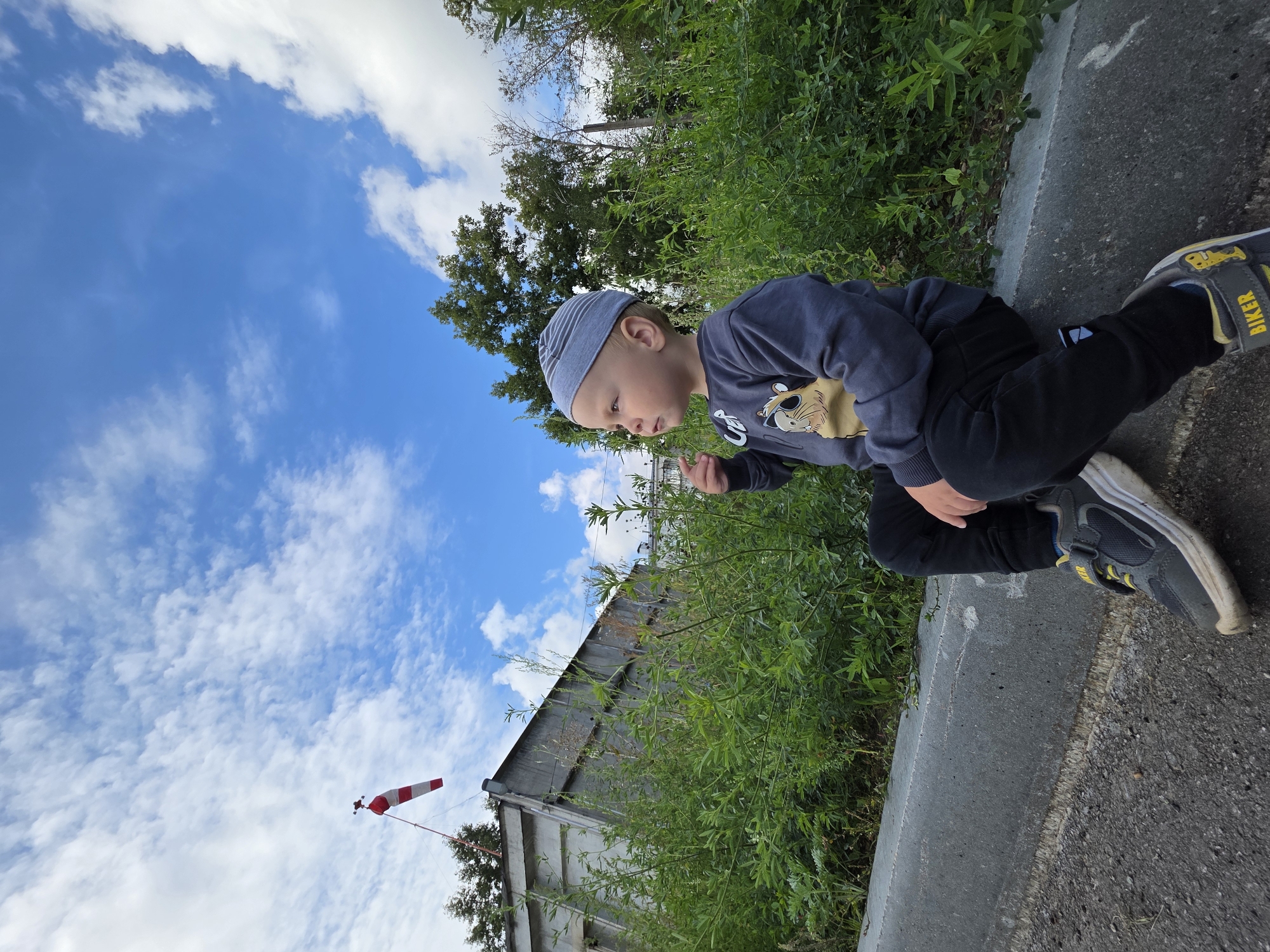 A young boy wearing a grey striped cap, a dark grey long-sleeved shirt with a cartoon image, black pants, and grey sneakers with yellow laces, sits on a concrete curb. He is looking to his left with his right hand slightly raised. The background features lush green vegetation, a grey metal structure, and a red and white windsock against a blue sky with white clouds. The scene appears to be outdoors, possibly near an airfield or industrial area in the city of Chaikovsky, Russia.