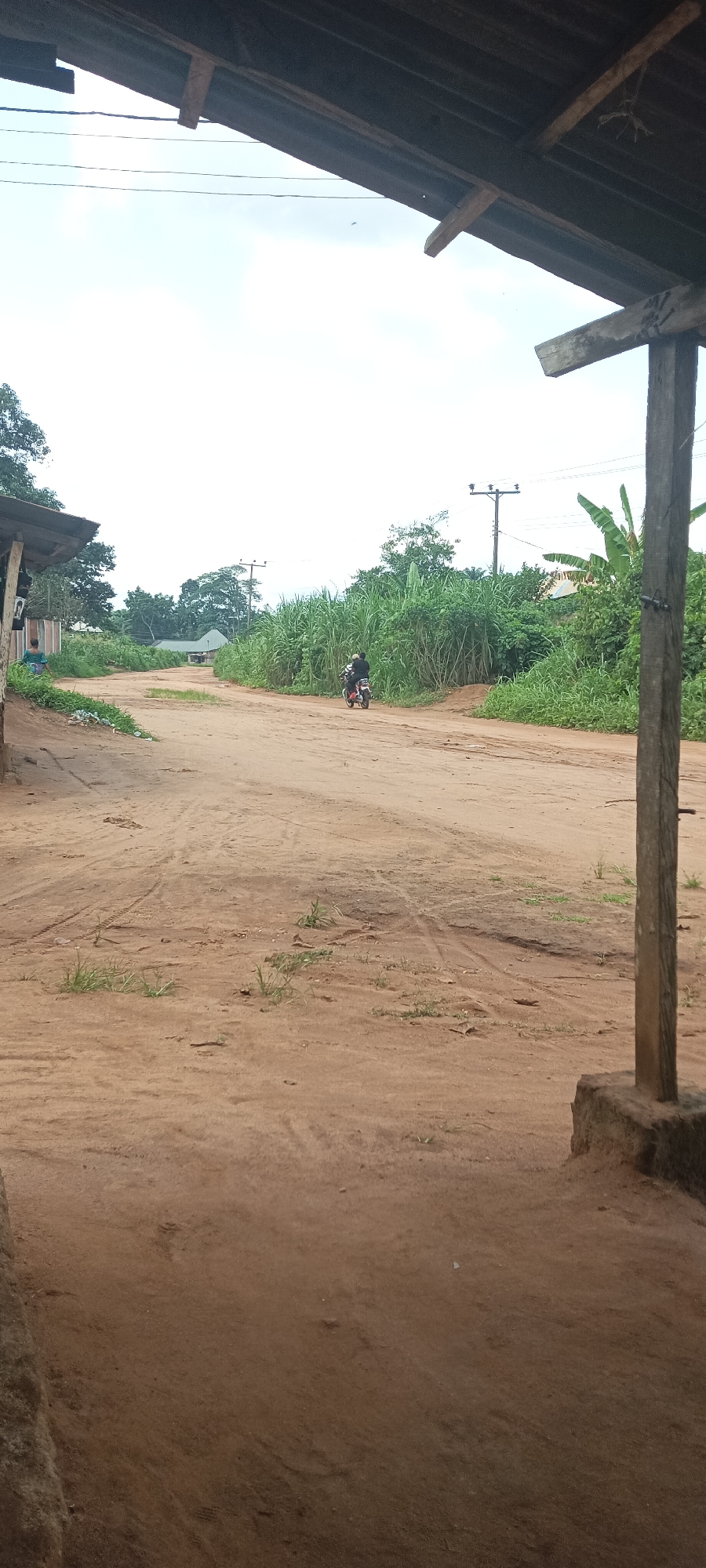 The scene is an unpaved road in Amauzu, Nigeria. The road is brown and sandy with tire tracks. On the road, a motorcycle with two people rides towards the viewer's perspective. The road is bordered by green vegetation and trees. Overhead power lines cross the sky. The image is taken from under a wooden structure, likely a shelter or building overhang. A wooden post is visible on the right side of the frame.
