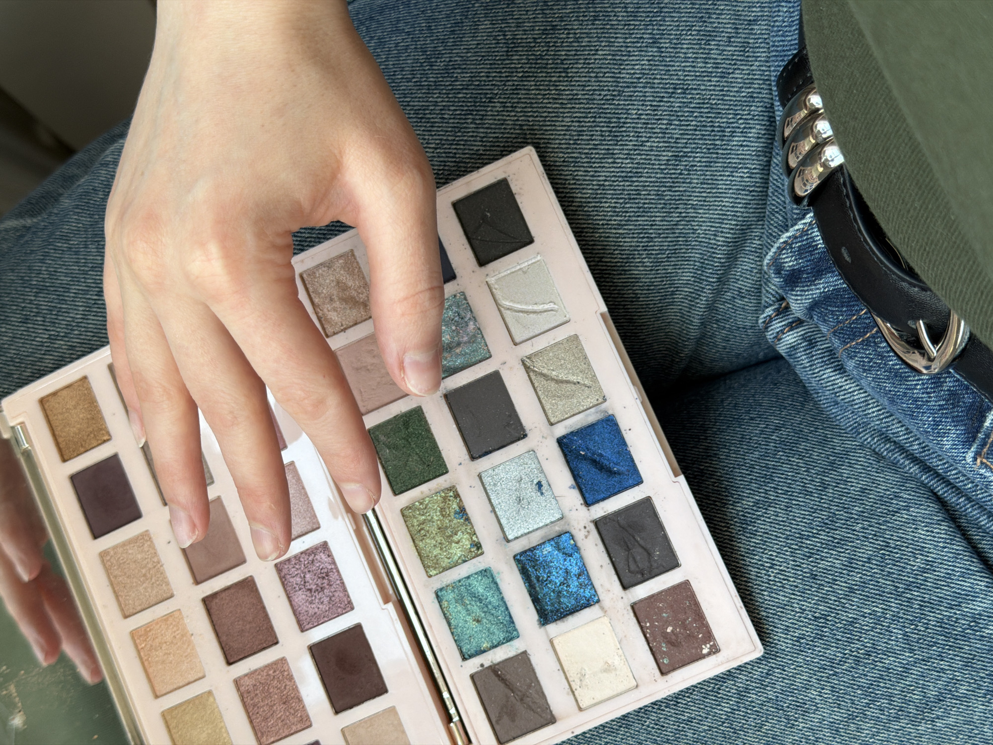 A person's hand rests on an open eyeshadow palette. The palette is predominantly pink and contains a variety of eyeshadow colors, including browns, greens, and blues. The palette is resting on a lap covered in blue denim jeans. A black belt with silver hardware is also visible. The location is not discernible.