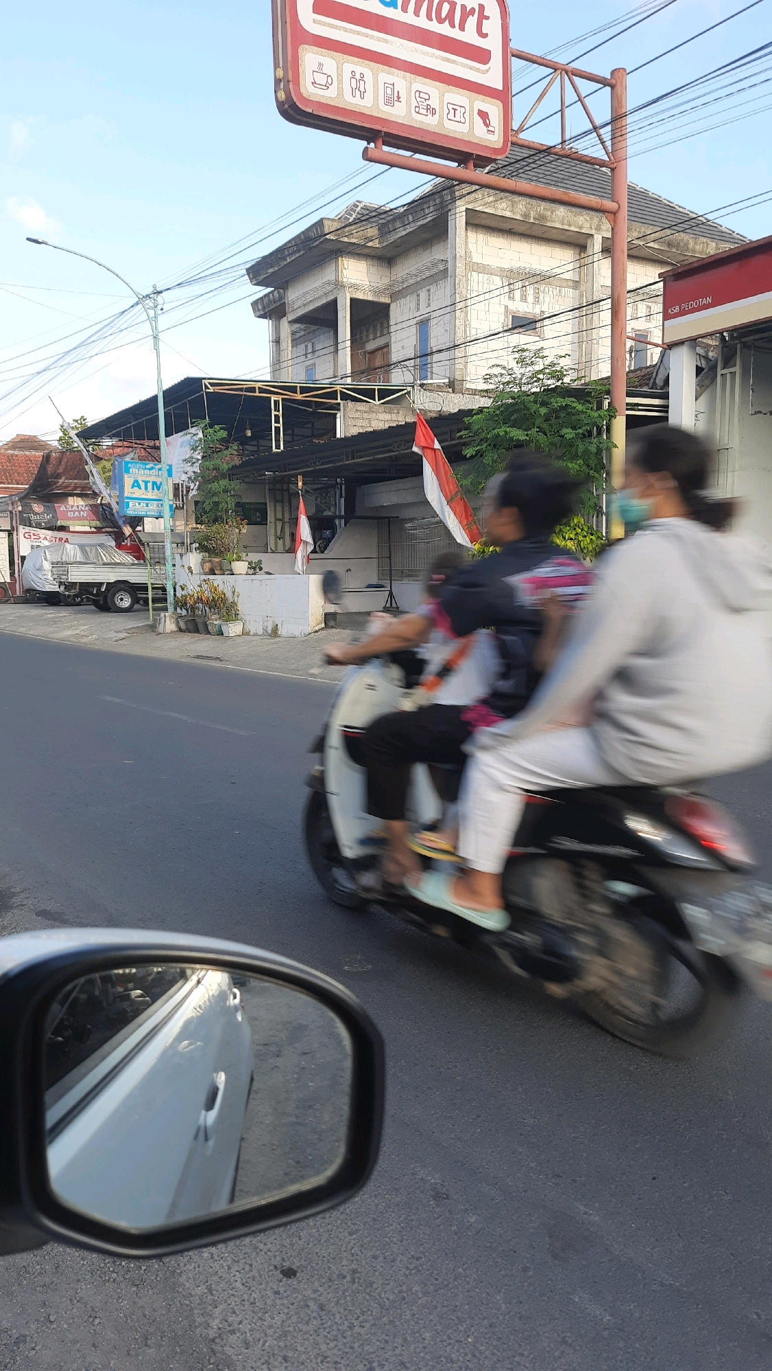 The image depicts a street scene in Bangorejo, Indonesia, on a clear day with bright sunlight. In the foreground, the side mirror of a white car is visible, reflecting the road and the car's side panel. The primary focus is a motorcycle traveling on the road, carrying two people who are blurred due to motion. The rider, wearing a black jacket, is in front, and a passenger, wearing a light-colored hoodie and a light blue mask, is behind. Both are wearing casual clothing and are partially visible. The motorcycle is black with white accents.
In the background, a two-story building under construction, with exposed brickwork and scaffolding, dominates the scene. A sign for "Alfamart," a convenience store, is prominently displayed with its red and white branding. Another sign reads "AGEN MANDIRI ATM." Indonesian flags are visible, one hanging from the building and another smaller one attached to a pole. The street is paved and appears to be a public road. To the left, a white pickup truck is parked, partially covered with a tarp. The overall environment suggests a typical urban or suburban setting in Indonesia, with a mix of ongoing construction and established businesses.