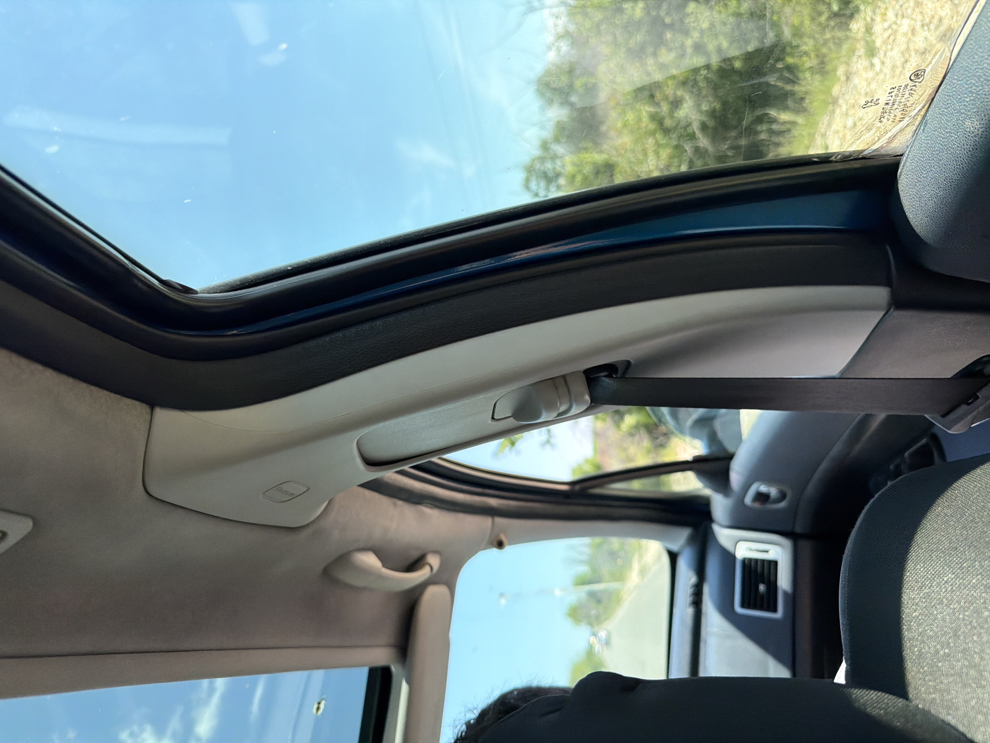The image shows the interior of a car, specifically the roof and part of the dashboard. The car has a sunroof. Outside the car, vegetation can be seen. The sky is blue and clear. The location is likely outdoors, as evidenced by the natural light and vegetation. There are no discernible people or actions taking place, beyond the implicit fact of someone being in the car. The location of Belovitsa, Bulgaria is not evident from the image.