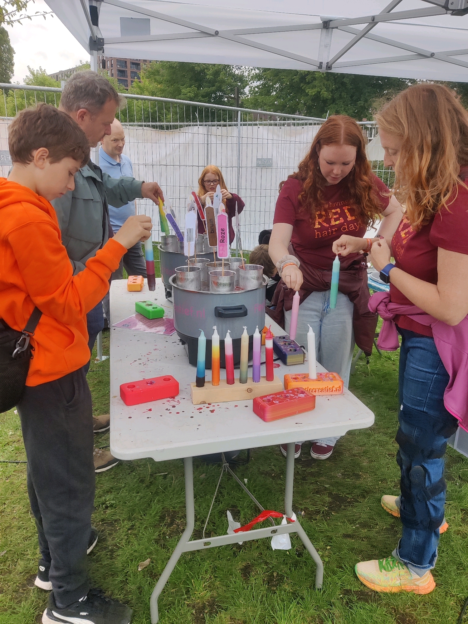 The image depicts an outdoor scene, likely a market or event, in Tilburg, Netherlands. In the foreground, a young boy wearing an orange hoodie and black pants stands at a white folding table. He is engaged in a candle-making activity, holding a partially dipped candle. To his right, a woman with red hair, wearing a maroon t-shirt that reads "having RED hair day," is also participating, holding a light blue candle. Another woman, also with red hair and wearing a similar maroon t-shirt, is to her right, holding a pink candle. Behind the boy, a man in a grey jacket and a man in a blue shirt are observing the activity. The table is laden with various objects related to candle making, including molds, colored wax sticks, and a large pot. In the background, a white tent structure provides shade, and a metal fence separates the area from more distant trees and buildings. The ground is covered with grass and appears to be outdoors. The overall atmosphere suggests a hands-on craft event.